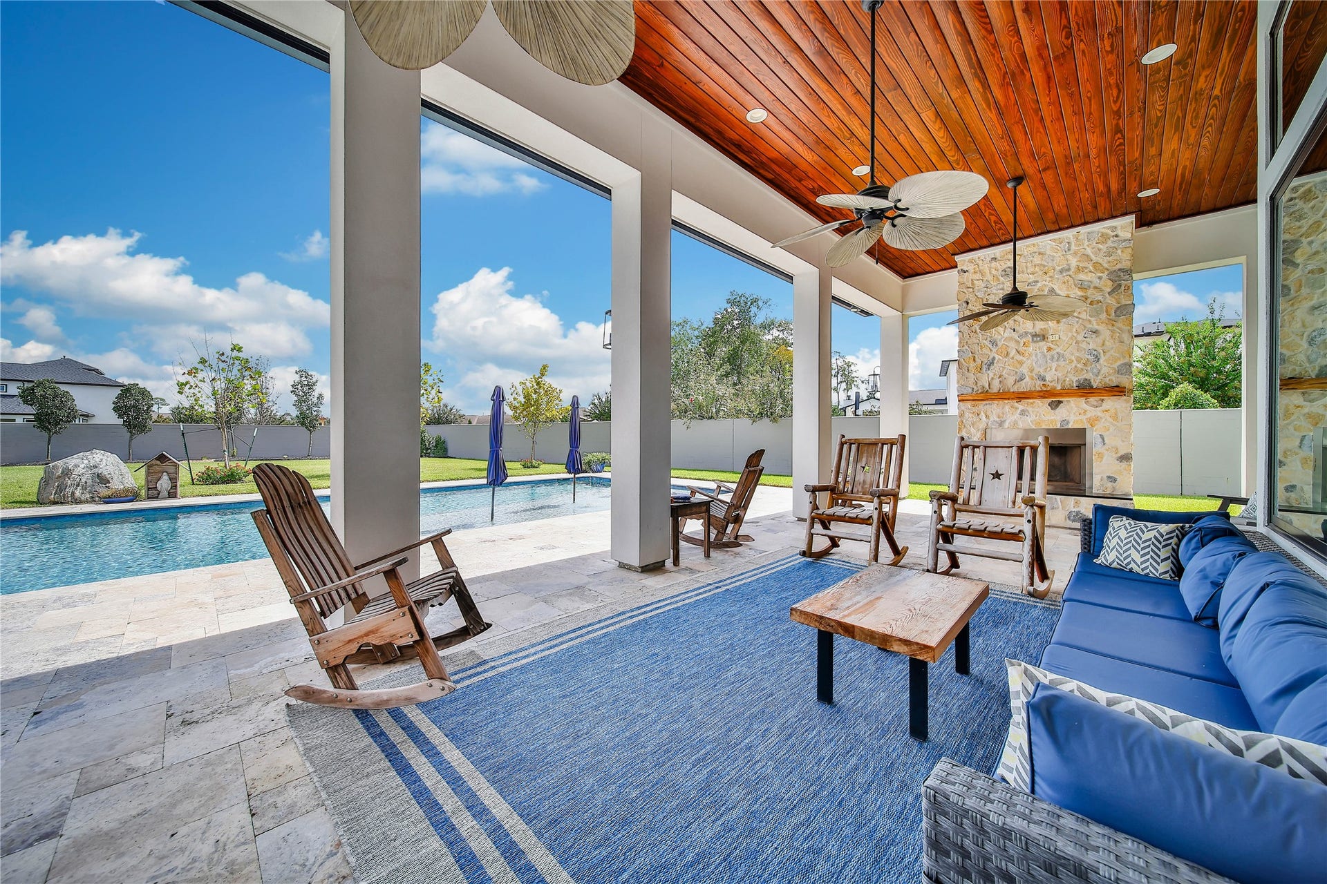 24718 Shadow Pond Drive, Spring, TX 77389 Elegant outdoor living space featuring a covered patio with wood ceiling, stone fireplace, and ceiling fans, overlooking a sparkling pool and landscaped yard.