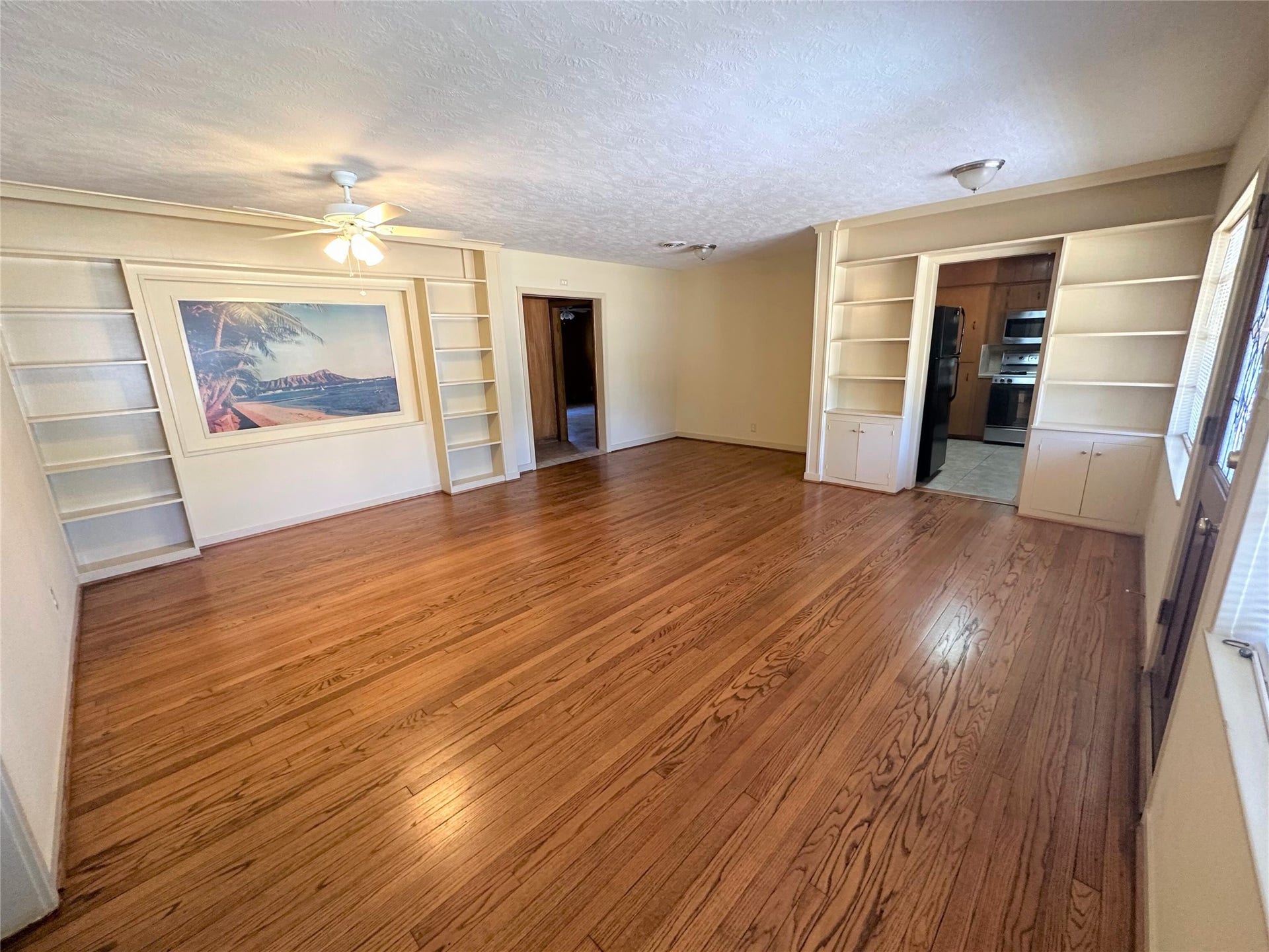 Another view of large living room with tons of built-ins, ceiling fan and beautiful hardwood floors.  Doorway in middle leads to primary suite hallway and additional living area/den.  Doorway on right leads to kitchen.