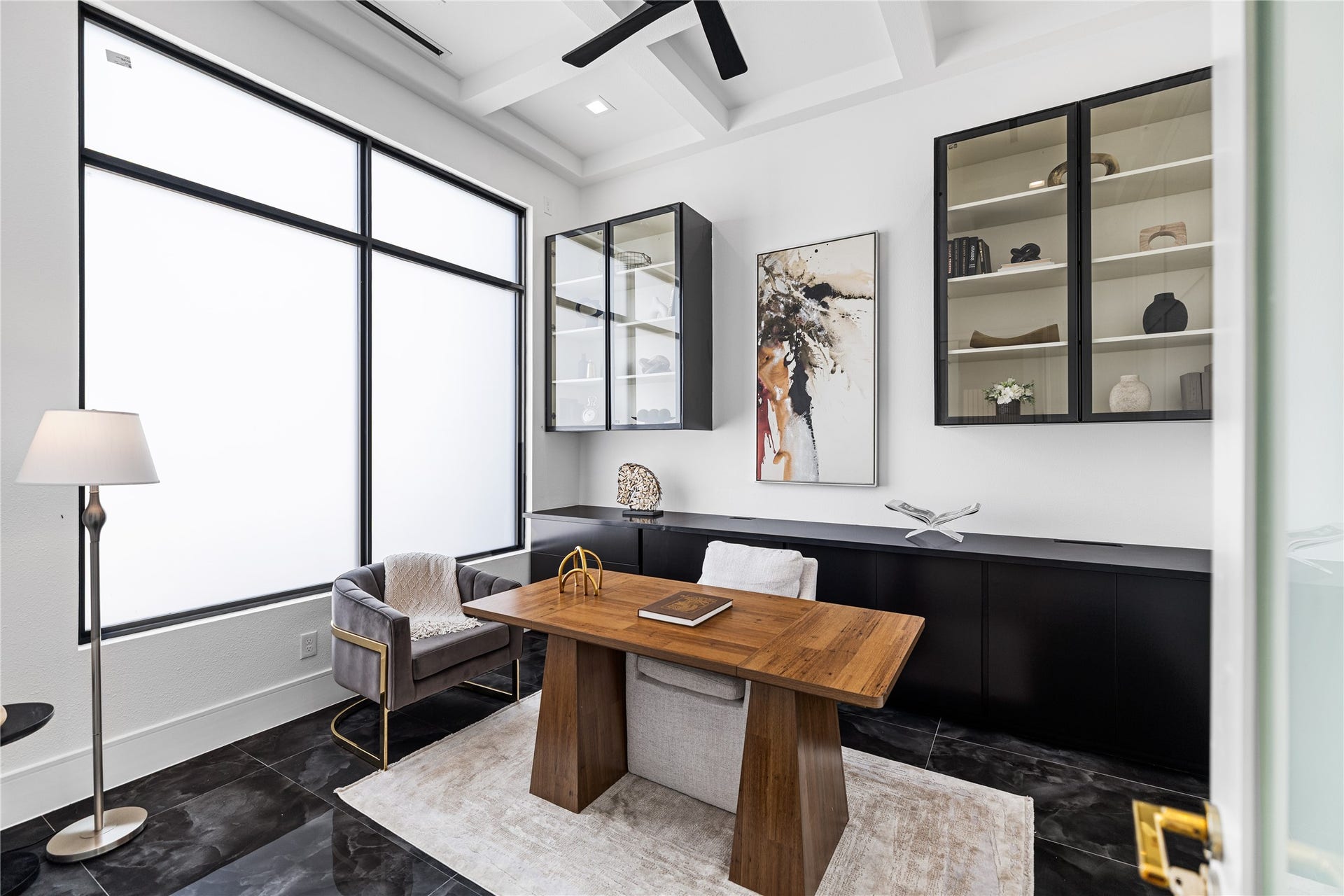A study/office with a striking black and white palette: black custom shelving along the lower walls against a white coffered ceiling. Large windows features smart frosted glass that switches to clear at the touch of a remote. Just a few of the sleek, high-tech privacy features.