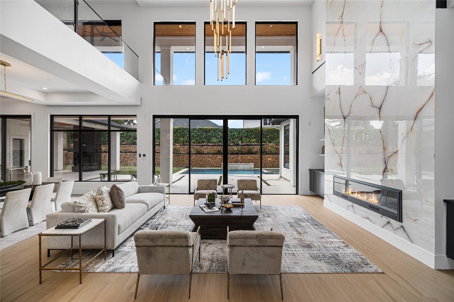 A dramatic two-story living room commands attention, anchored by a floor-to-ceiling marble fireplace that extends to the ceiling. Walls of glass frame the pool and outdoor views, flooding the space with natural light beneath a striking sculptural chandelier. This room is a rare blend of scale, warmth, and architectural presence.