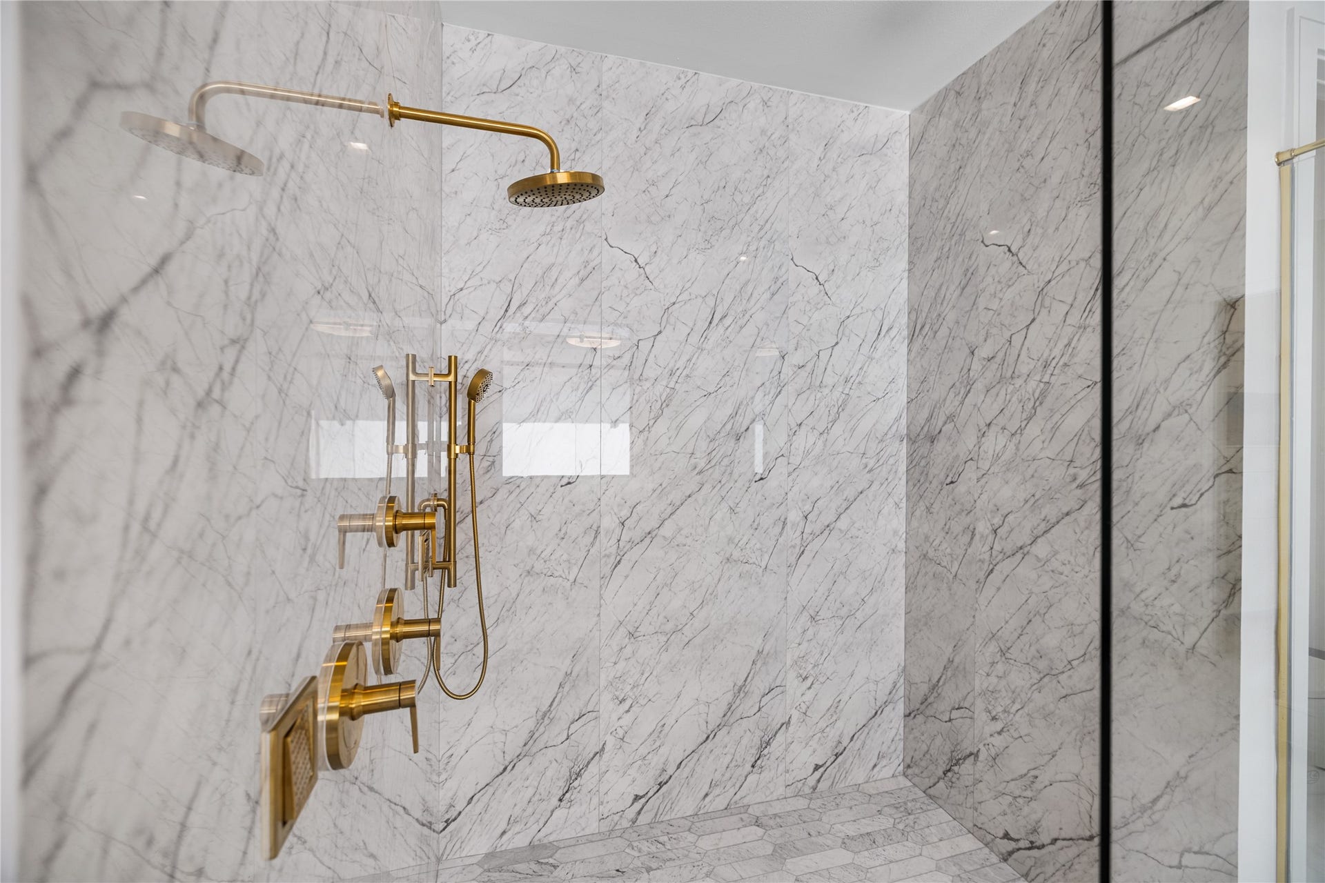 A closer look at the exquisite marble-clad primary walk-in shower, accented by elegant brass hardware and a spa-inspired shower head, handheld wand, and invigorating body sprays.