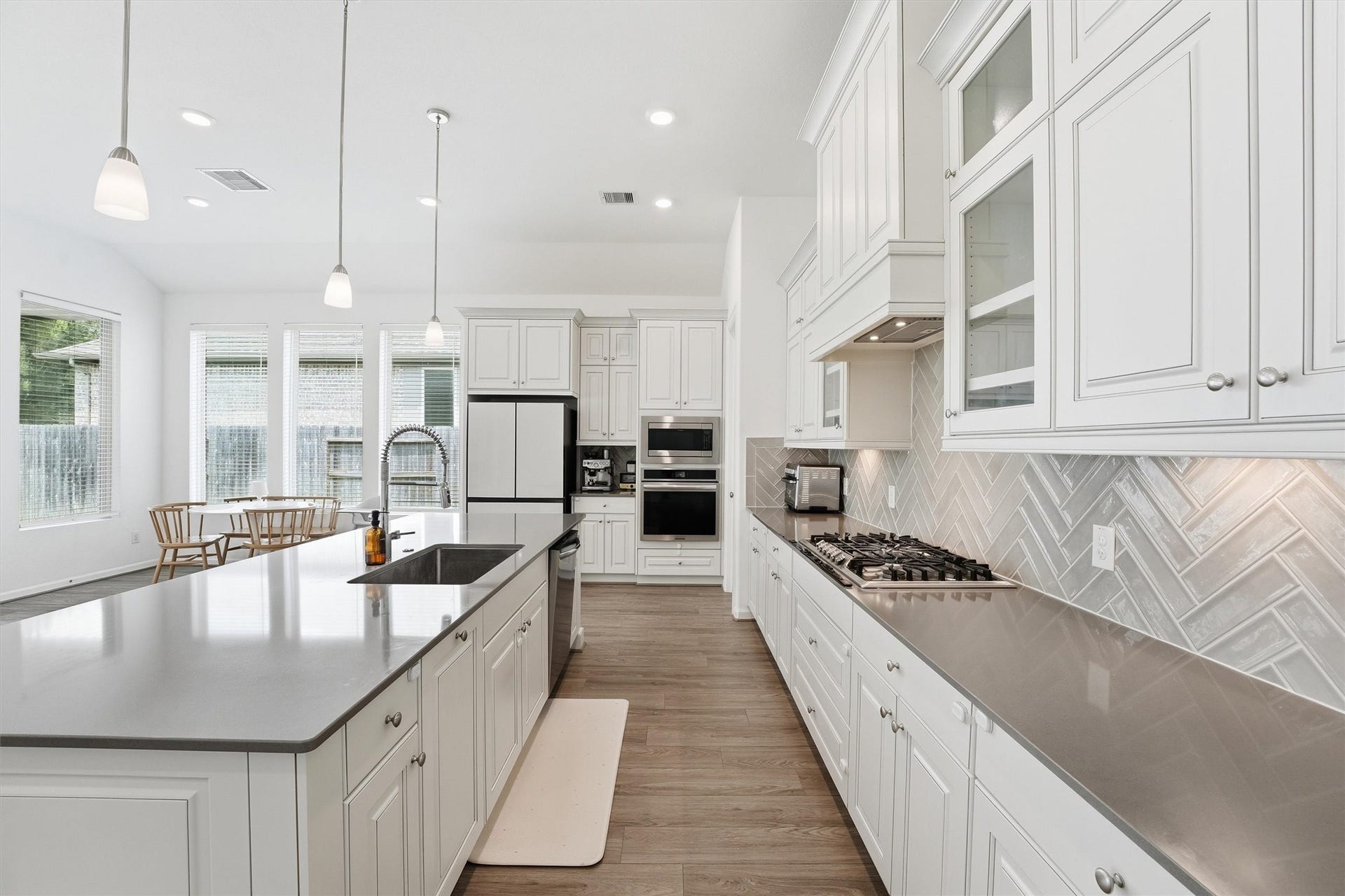 Spacious kitchen featuring sleek countertops, elegant white cabinetry, and a stylish herringbone backsplash. The large island with a sink and modern pendant lighting offers ample workspace, while the adjacent dining area is bathed in natural light from expansive windows.