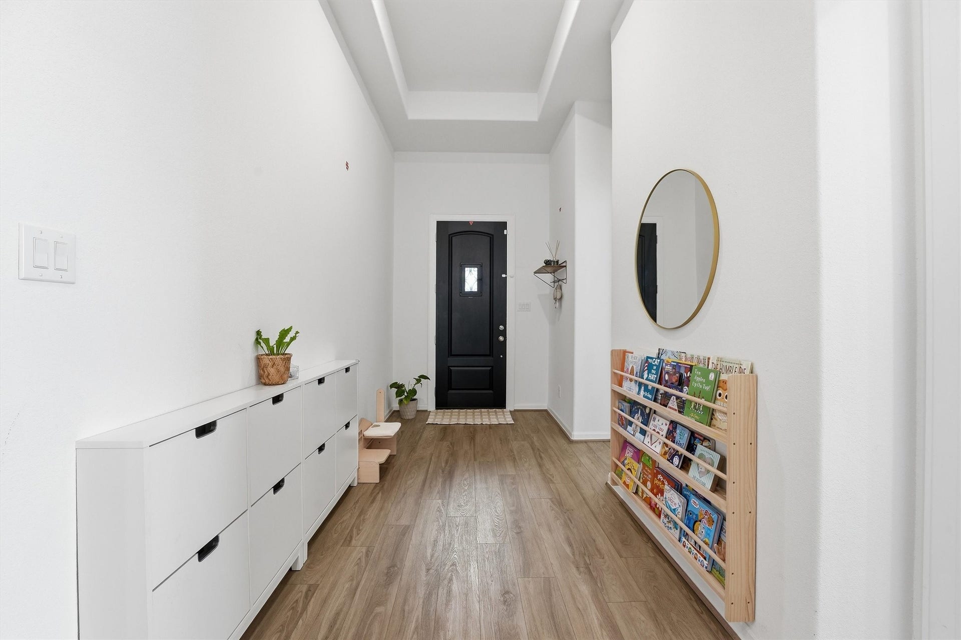 Bright and welcoming entryway featuring sleek wood-look flooring, a modern black door, and stylish storage solutions with a built-in book rack and shoe cabinet.