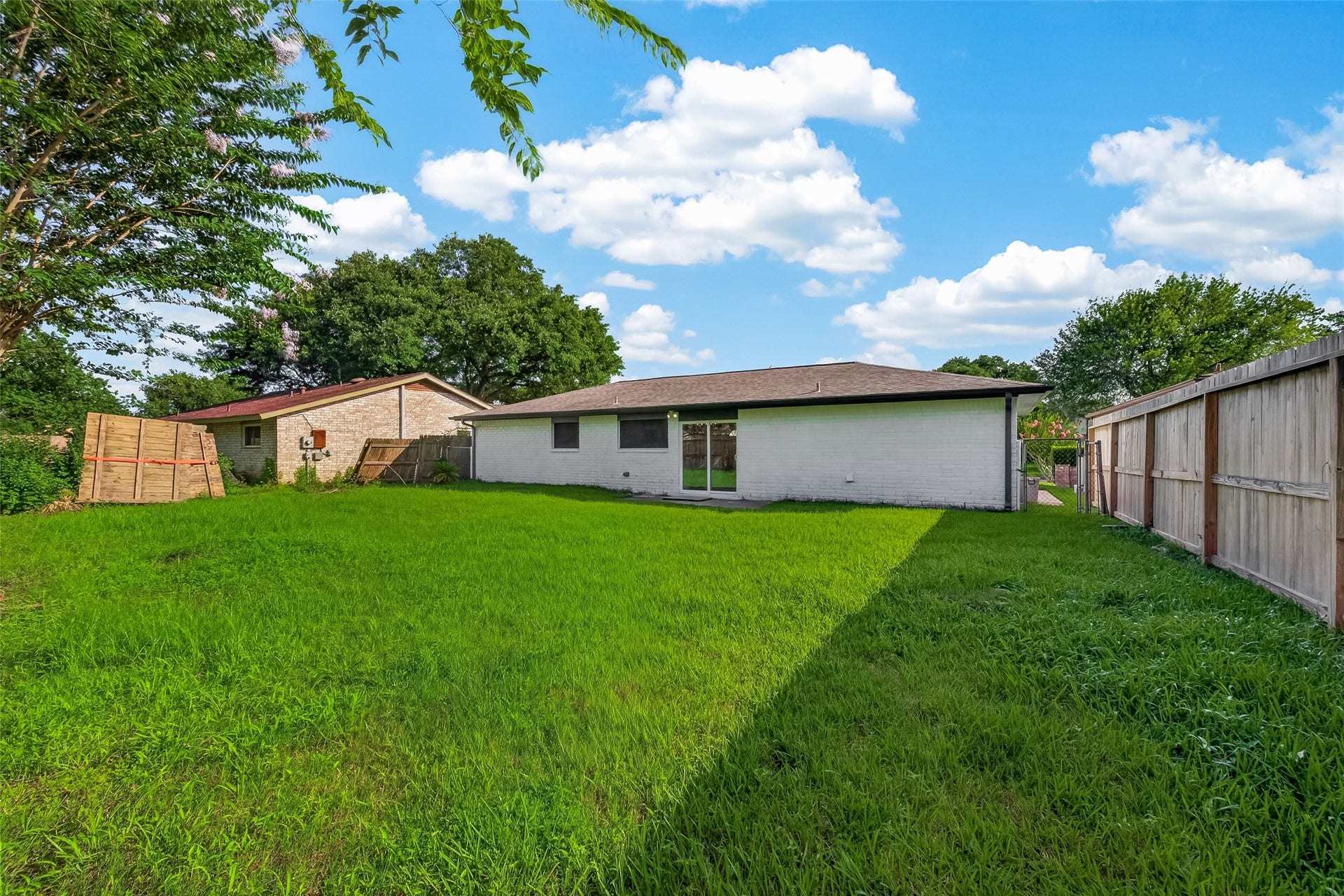 16619 Tibet Road, Friendswood, TX 77546 Spacious backyard with lush green grass, bordered by a wooden fence for privacy. The rear of the house is visible, featuring a simple, single-story design with large windows. Ideal for outdoor activities and relaxation.