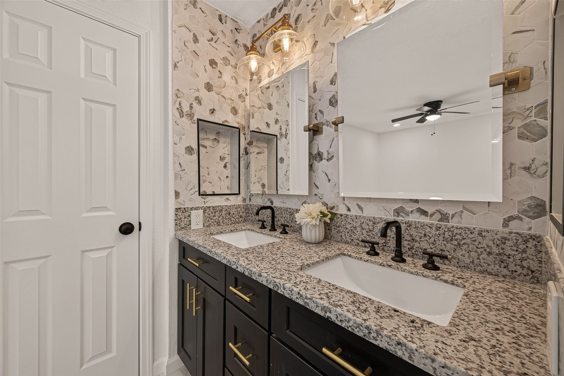 16619 Tibet Road, Friendswood, TX 77546 Modern bathroom with dual sinks, granite countertops, and dark cabinetry. Features stylish hexagonal tile backsplash, dual mirrors, and elegant lighting.