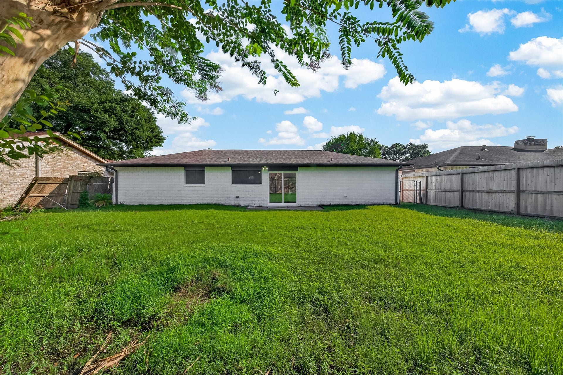 16619 Tibet Road, Friendswood, TX 77546 This photo shows a spacious backyard with a well-maintained lawn, bordered by a wooden fence. The back of the house has a simple, white exterior with a central sliding glass door, providing easy access to the yard. A tree in the foreground adds a touch of greenery.