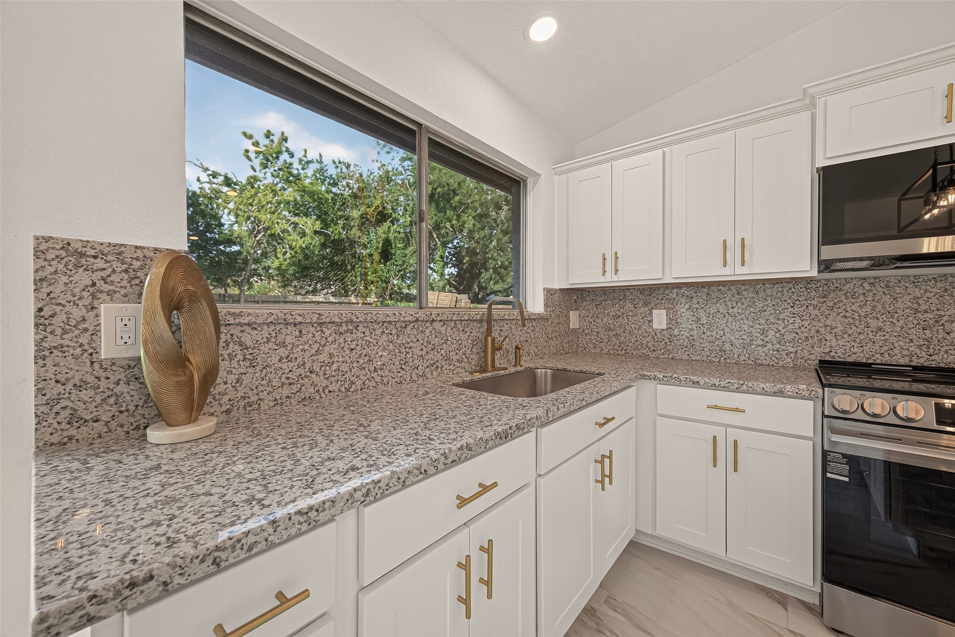 16619 Tibet Road, Friendswood, TX 77546 This modern kitchen features sleek granite countertops, white cabinetry with gold hardware, a stainless steel sink, and a large window offering plenty of natural light.