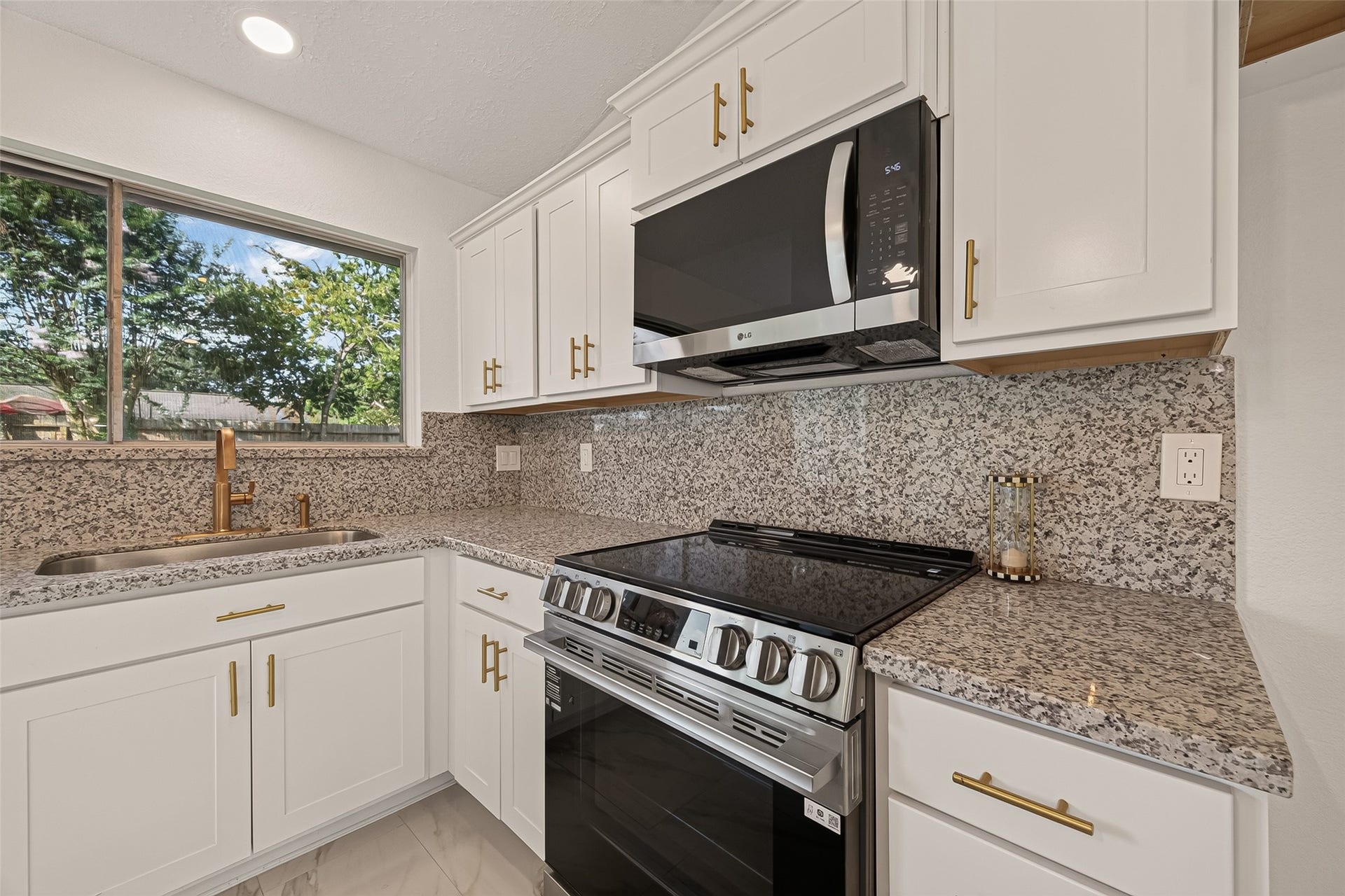 16619 Tibet Road, Friendswood, TX 77546 This modern kitchen features sleek white cabinetry, granite countertops, and stainless steel appliances, including a built-in microwave and stove. A large window provides natural light, and there are stylish gold fixtures and handles.