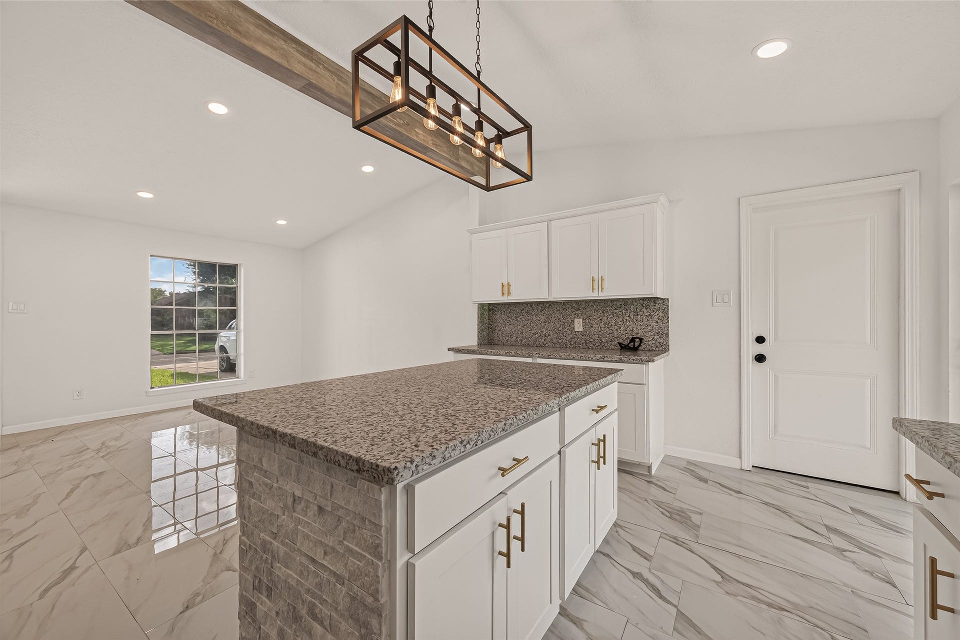 16619 Tibet Road, Friendswood, TX 77546 This photo showcases a modern kitchen with sleek granite countertops, white cabinetry, and stylish gold hardware. The open layout features a central island with a brick accent, under a contemporary light fixture. The space is bright with natural light from a large window, complemented by polished tile flooring.