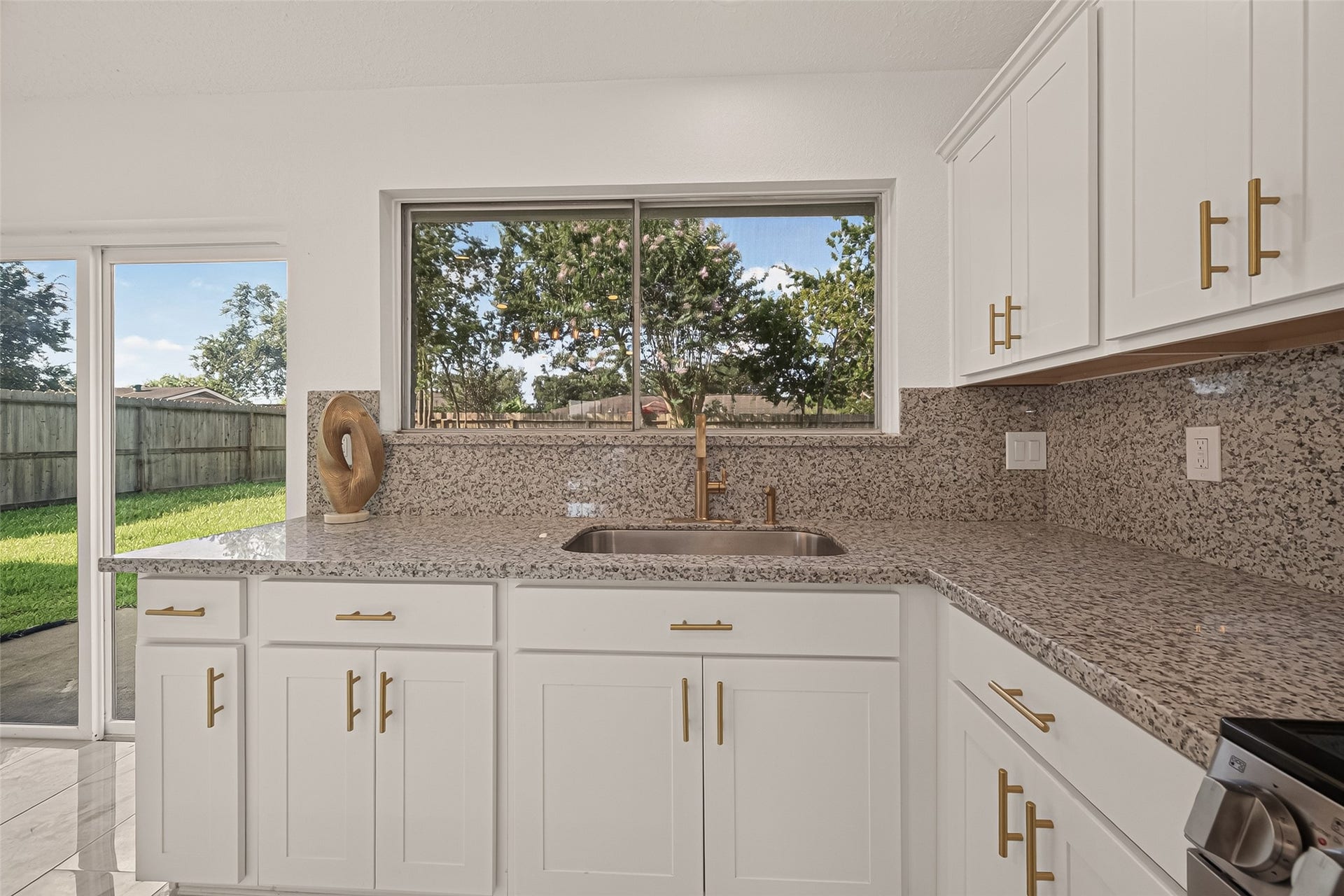 16619 Tibet Road, Friendswood, TX 77546 This kitchen features elegant granite countertops, white cabinetry with gold hardware, and a large window offering a view of the backyard. Natural light fills the space, creating a bright and inviting atmosphere.