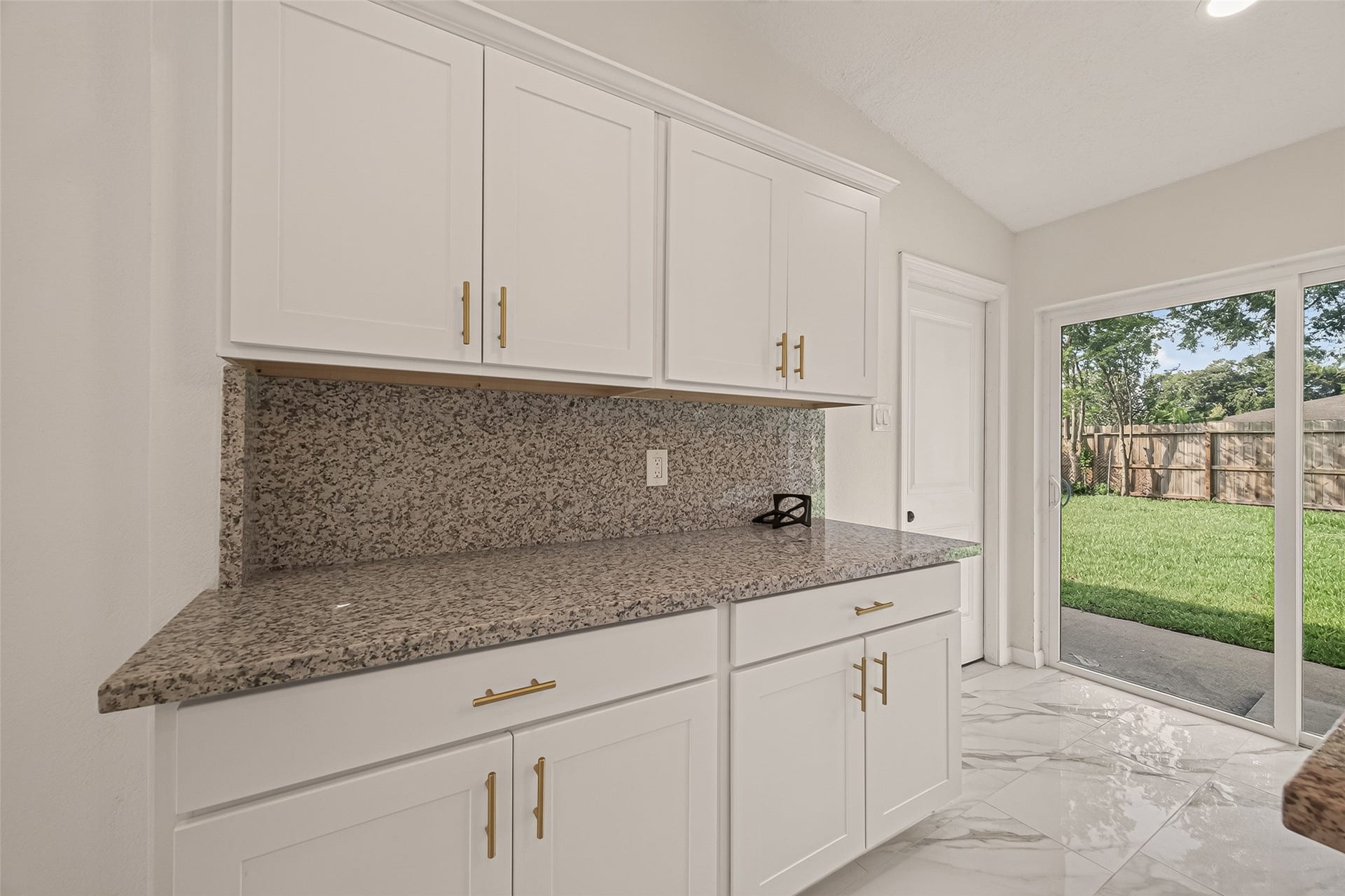 16619 Tibet Road, Friendswood, TX 77546 This photo showcases a bright kitchen space featuring elegant white cabinets with modern hardware, a speckled granite countertop, and a large sliding glass door opening to a lush, fenced backyard. The room is highlighted by glossy, tiled flooring.