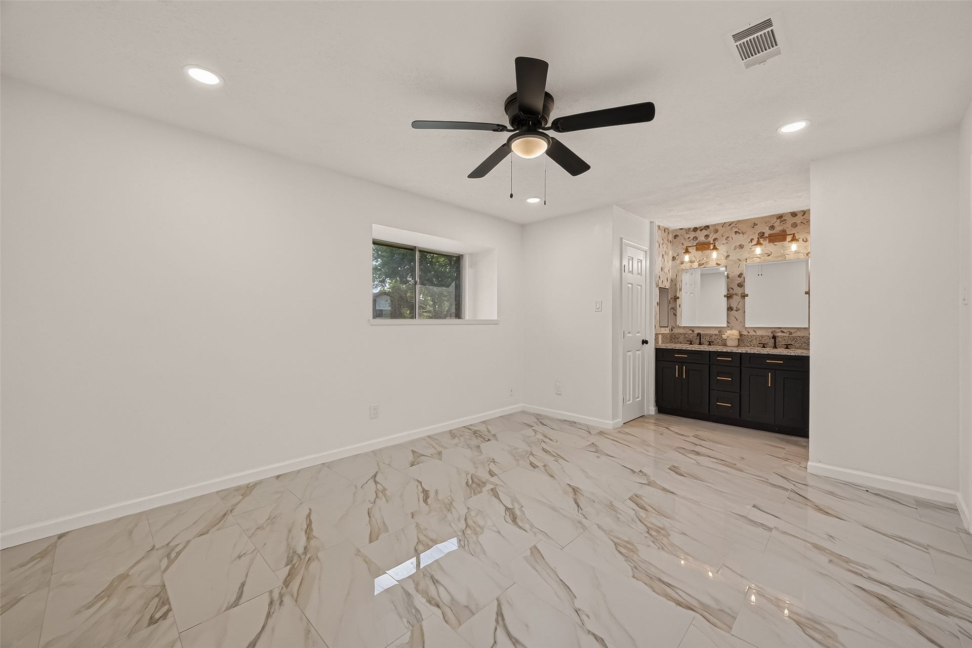 16619 Tibet Road, Friendswood, TX 77546 This photo showcases a modern bedroom with glossy marble-style tile flooring, a ceiling fan, and recessed lighting. It has a small window for natural light and an open ensuite bathroom featuring a dual-sink vanity with dark cabinetry and decorative lighting.
