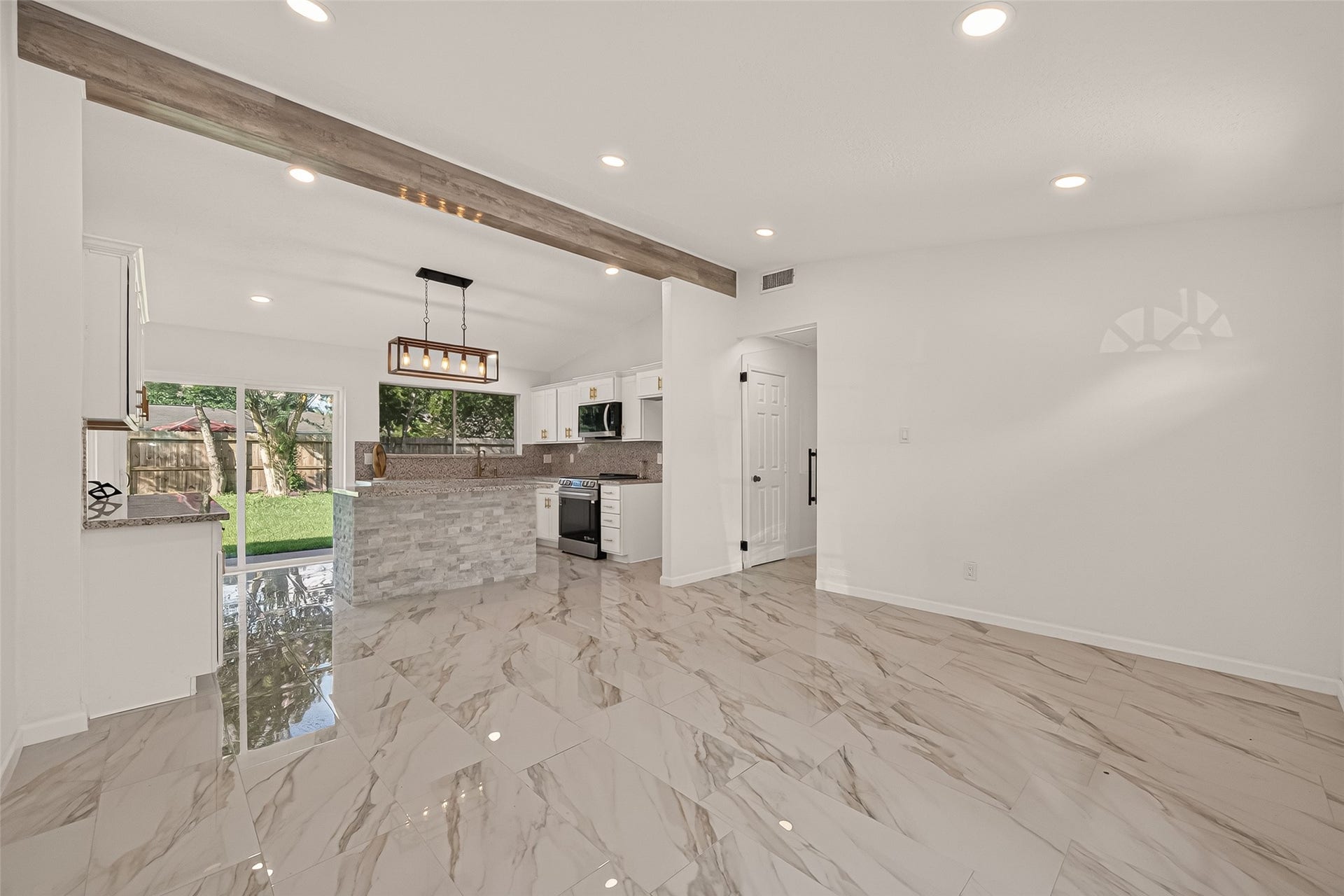 16619 Tibet Road, Friendswood, TX 77546 This photo showcases a modern open-concept kitchen and living space with elegant marble tile flooring, recessed lighting, and a stylish wood beam accent. The kitchen features a central island with stone detailing, contemporary light fixtures, and large windows offering plenty of natural light and views of the backyard.