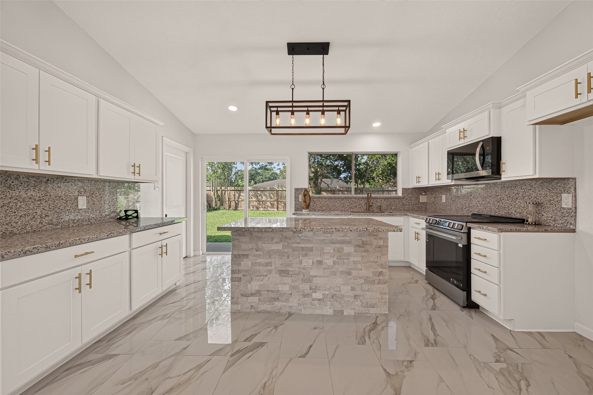16619 Tibet Road, Friendswood, TX 77546 This modern kitchen features sleek white cabinetry with gold handles, granite countertops, and a central island with stone accents. It boasts stainless steel appliances, a stylish rectangular pendant light, and large windows allowing natural light to fill the space, complemented by polished marble-look tile flooring.