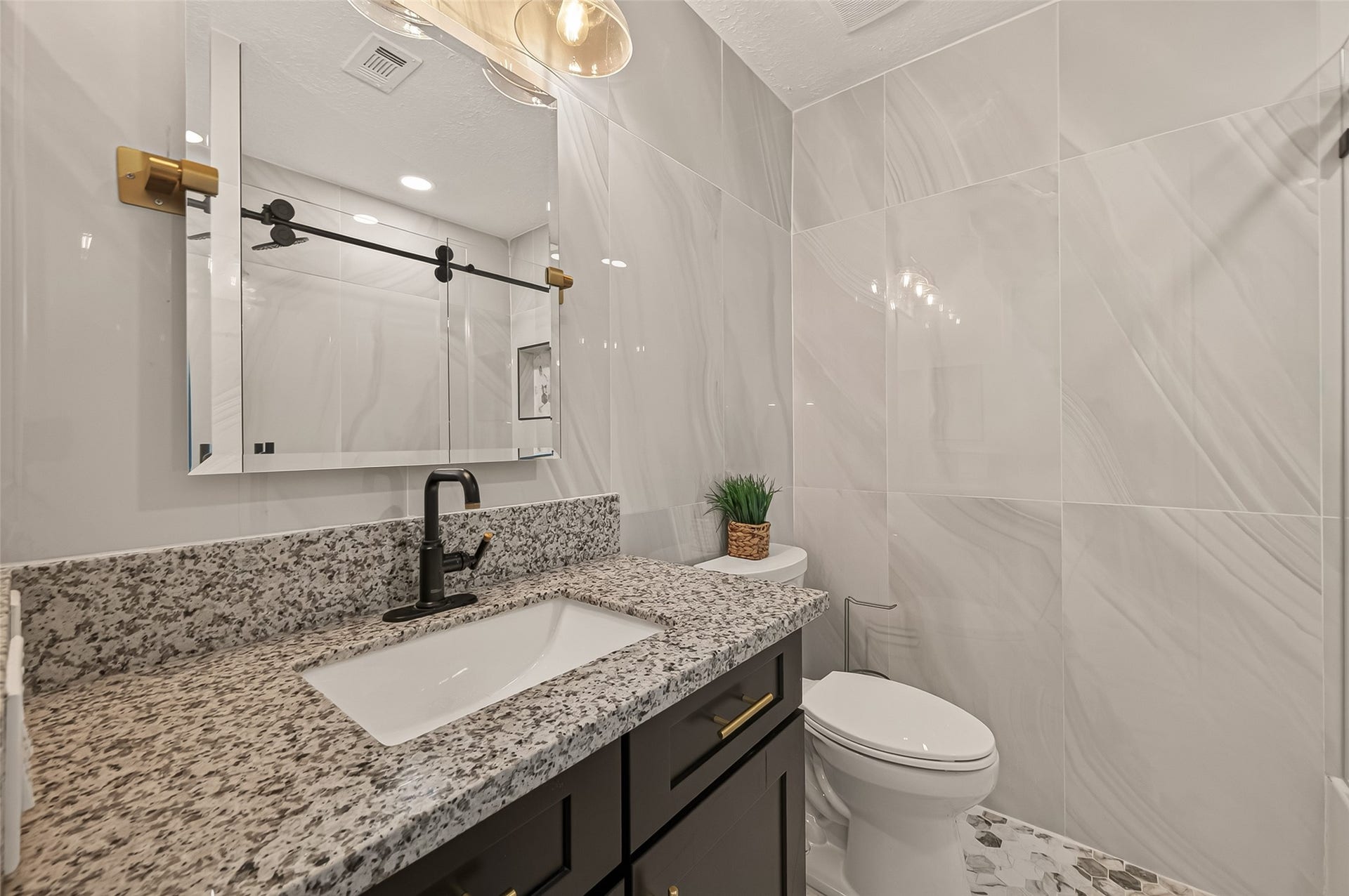 16619 Tibet Road, Friendswood, TX 77546 This modern bathroom features a sleek design with large marble-style wall tiles, a granite countertop, and a black faucet. The vanity has ample storage with dark cabinets and gold hardware. A stylish mirror and elegant lighting add a touch of sophistication.