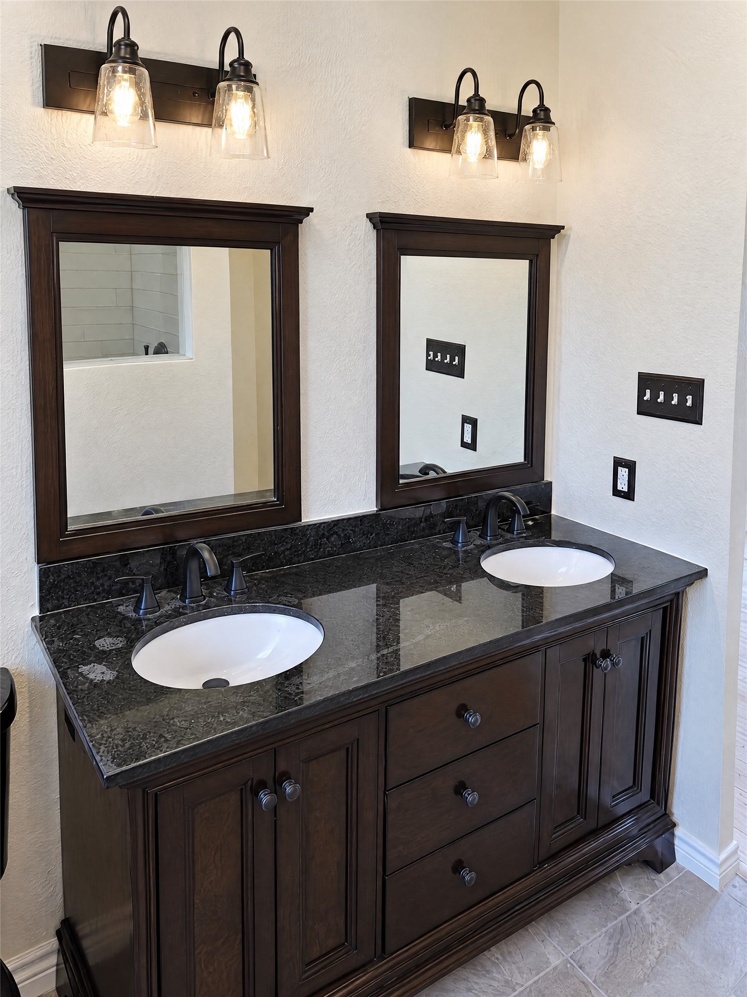 16411 Amcreek Road, Houston, TX 77068 Step into the en-suite bathroom showcasing an elegant dark wood double vanity topped with polished granite, matching framed mirrors, oil-rubbed bronze fixtures, and warm designer lighting.