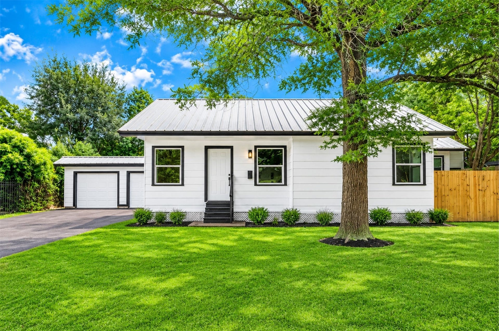 Charming white cottage with a metal roof, nestled under a large tree, featuring a detached two-car garage and a spacious, well-maintained yard.