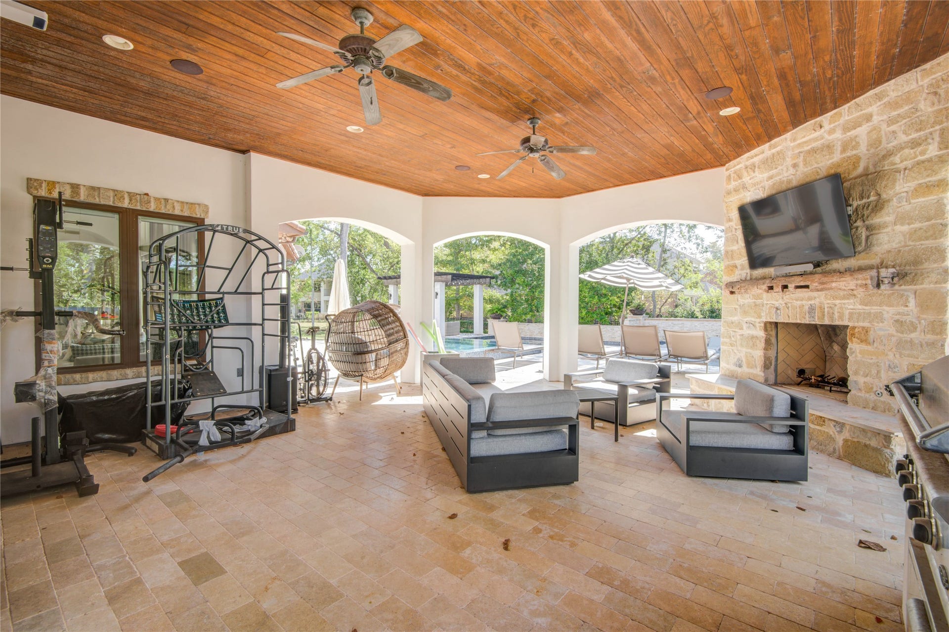 10 Sullivans Lane, Missouri City, TX 77459 Spacious outdoor patio with a cozy seating area includes a stone fireplace, wall-mounted TV, and ceiling fans. Large arched openings offer a view of the backyard, enhancing the indoor-outdoor living experience.
