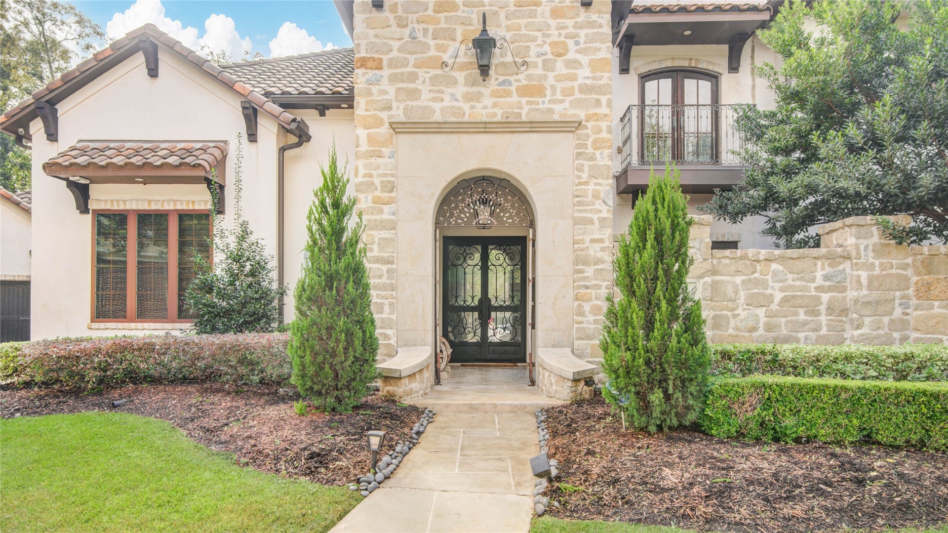 10 Sullivans Lane, Missouri City, TX 77459 This welcoming grand arched entry is framed by elegant stonework and tall, manicured evergreens. The walkway leads through beautifully maintained landscaping, highlighting the homes sophisticated European-inspired architecture. Rich wood accents, detailed trim, and a charming balcony add character and warmth to the faade. The overall feel is luxurious, inviting, and impeccably designedsetting the tone for the refinement found throughout the property.