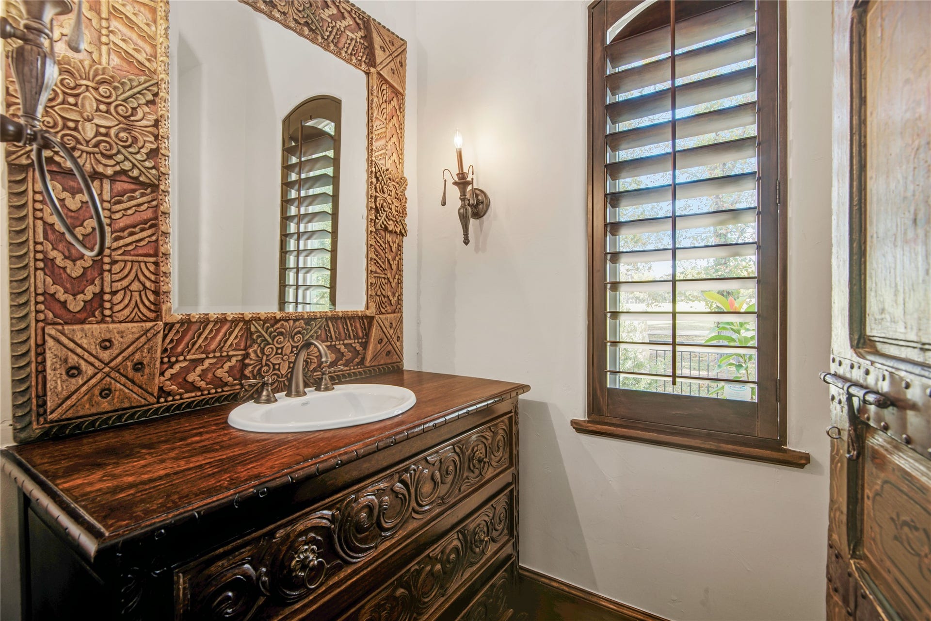 10 Sullivans Lane, Missouri City, TX 77459 This bathroom features a rustic design with a carved wooden vanity and an ornate mirror. Natural light filters through wooden plantation shutters, enhancing the warm, inviting atmosphere.