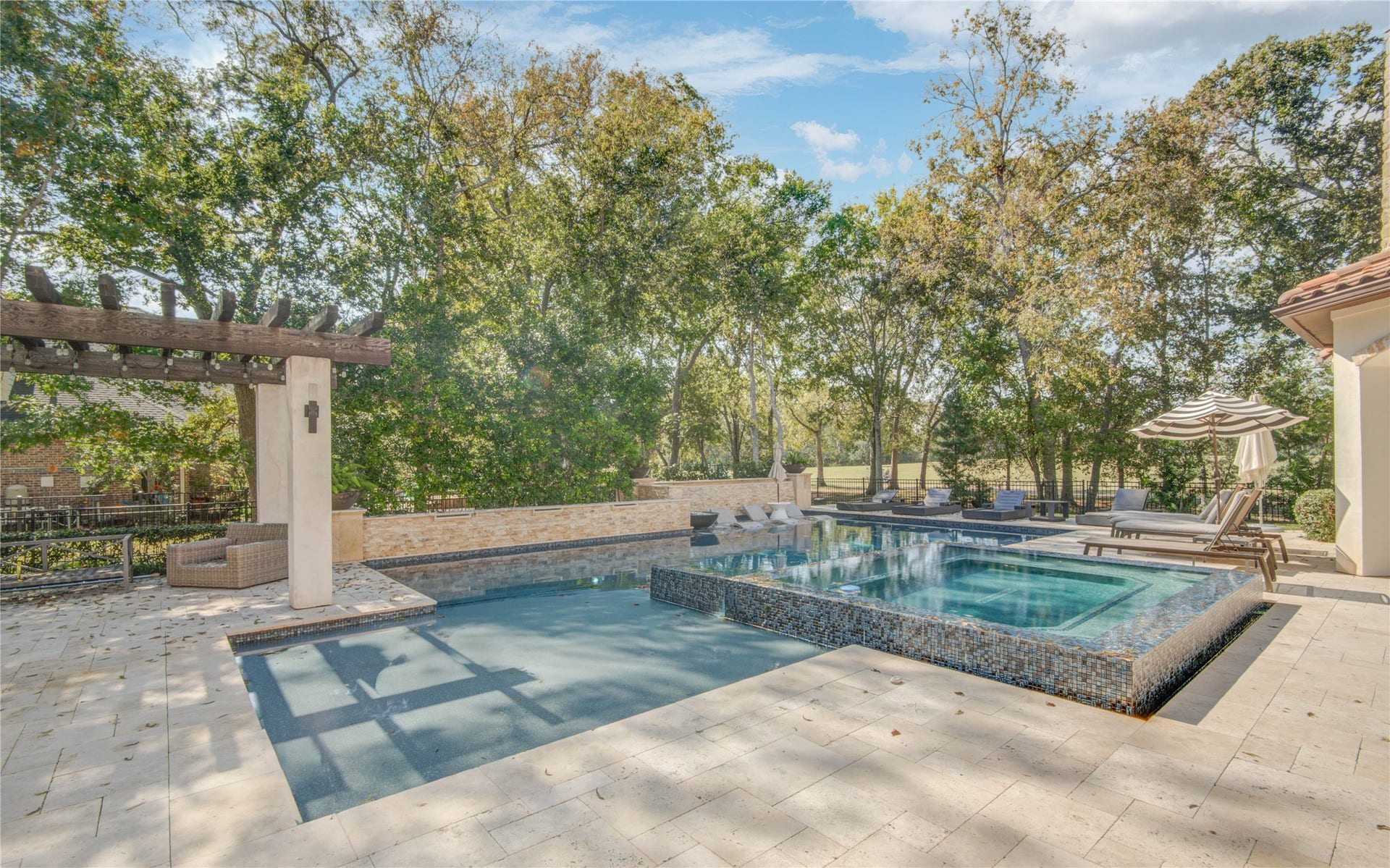 10 Sullivans Lane, Missouri City, TX 77459 Modern pool and hot tub, surrounded by elegant stone tile. There's a pergola with seating and a patio area all set against a backdrop of lush trees, offering privacy and tranquility.