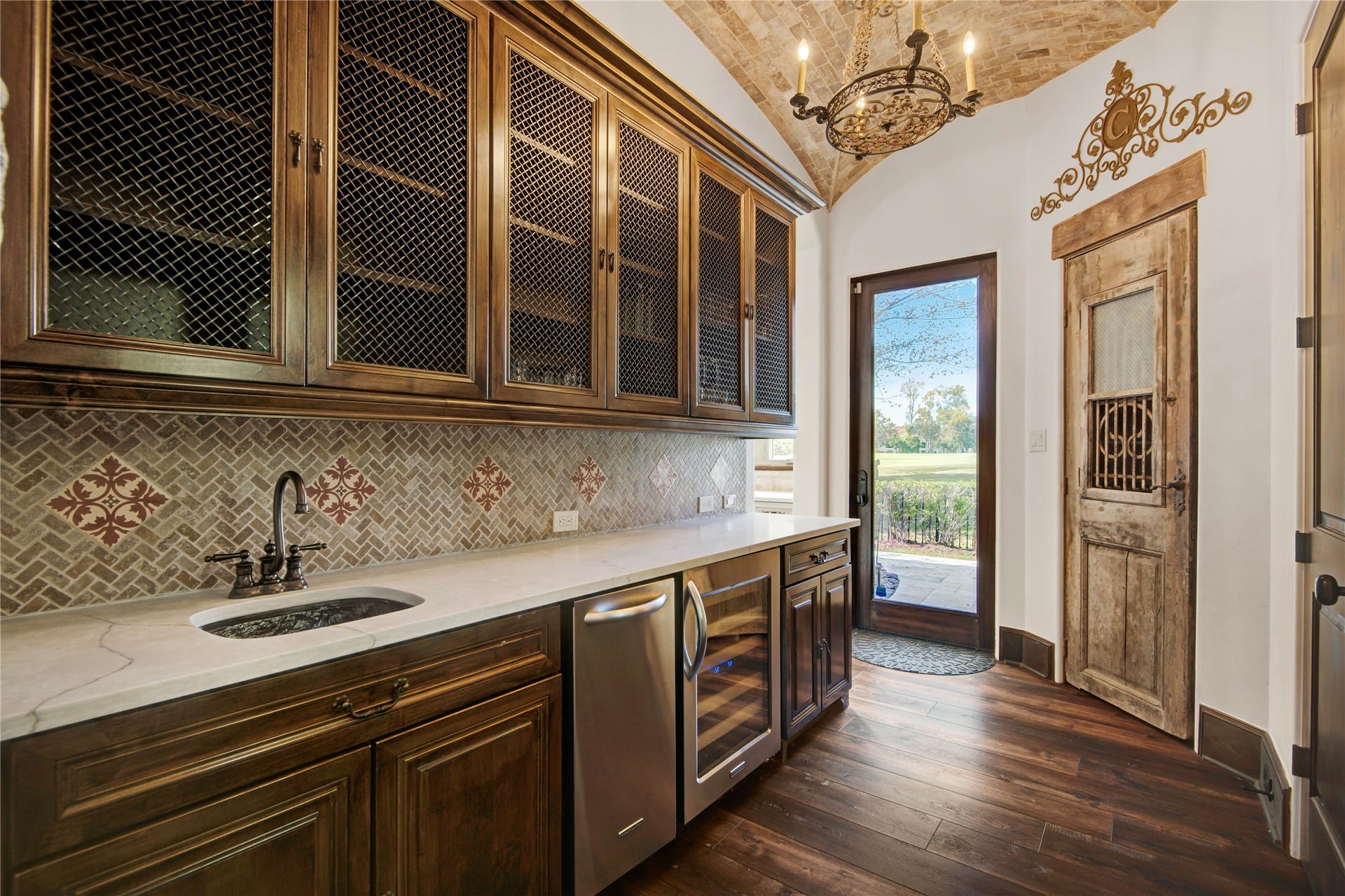 10 Sullivans Lane, Missouri City, TX 77459 This elegant butler's pantry features rich wooden cabinetry with mesh doors, a marble countertop, and a stylish tile backsplash. It includes a sink, ice maker, and wine fridge, with a rustic door and a decorative chandelier adding character. A glass door leads to an outdoor area with scenic views.