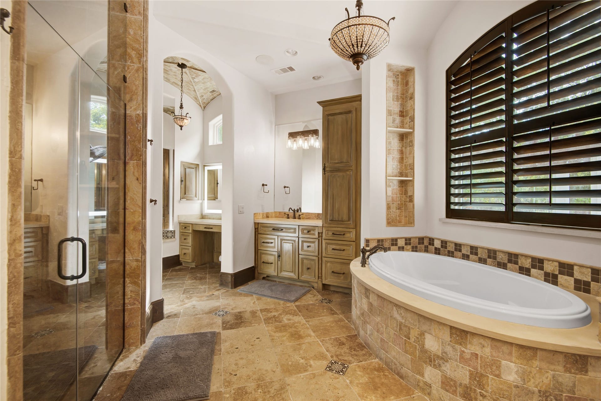 10 Sullivans Lane, Missouri City, TX 77459 Primary bathroom features elegant stone tiles, a spacious soaking tub, a large glass-enclosed shower, and dual sinks with ample cabinetry. Warm lighting and a window with wooden shutters add a cozy, yet luxurious feel.
