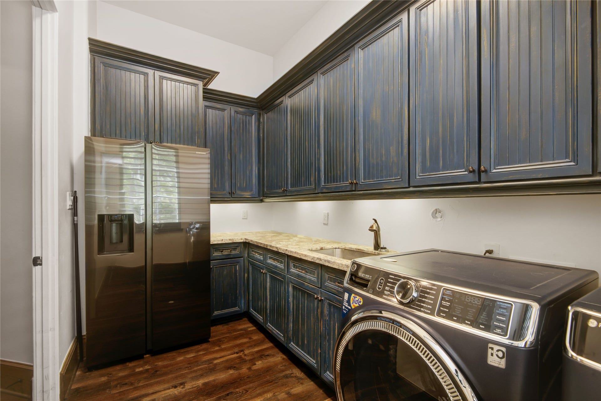 10 Sullivans Lane, Missouri City, TX 77459 Laundry room featuring dark wood flooring, blue paneled cabinets, granite countertops, and modern appliances, including a washer, dryer, and stainless steel refrigerator.