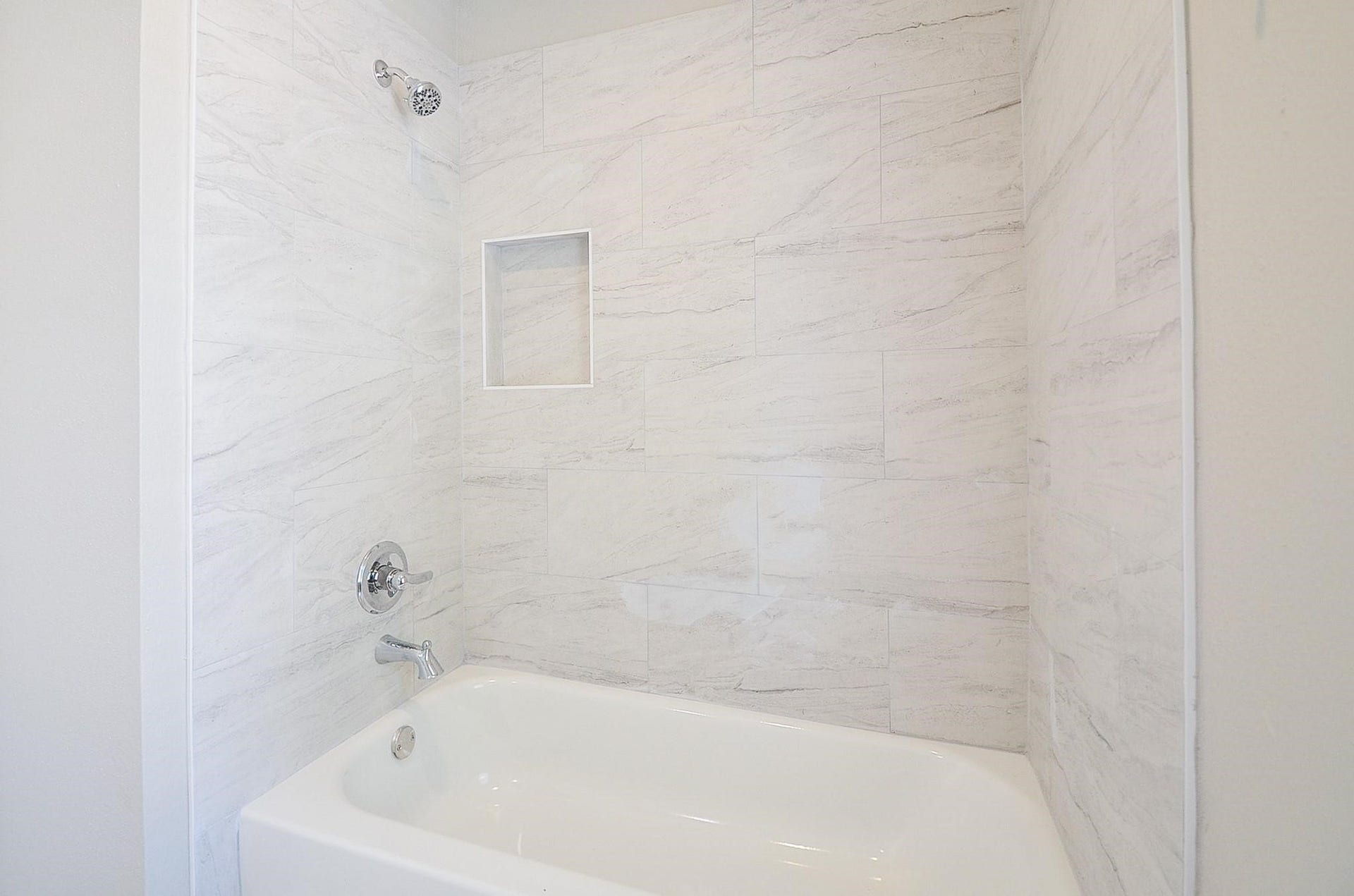 Discover an elegant bathroom oasis with a sleek white bathtub, surrounded by modern marble-patterned tiles and a handy built-in shelf for stylish storage.