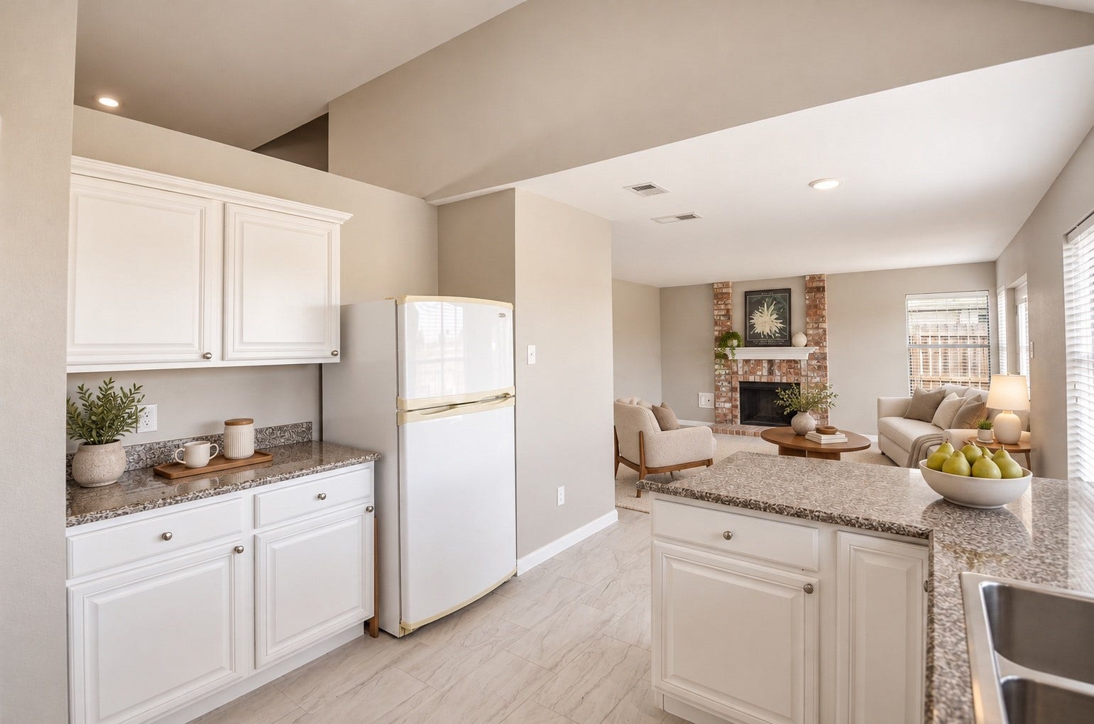 Bright and inviting kitchen with granite countertops and white cabinetry, seamlessly flowing into a cozy living room featuring a charming brick fireplace.