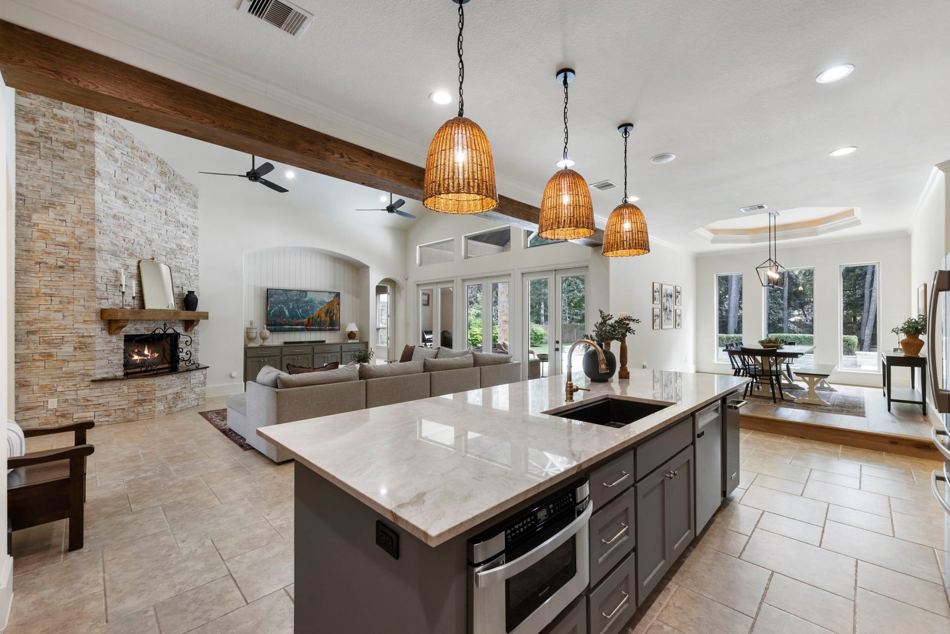 This vantage highlights the home's effortless flow, with the stacked-stone fireplace and sunlit breakfast nook visible from the expansive island. A rustic ceiling beam and tray-ceiling dining area add architectural interest.