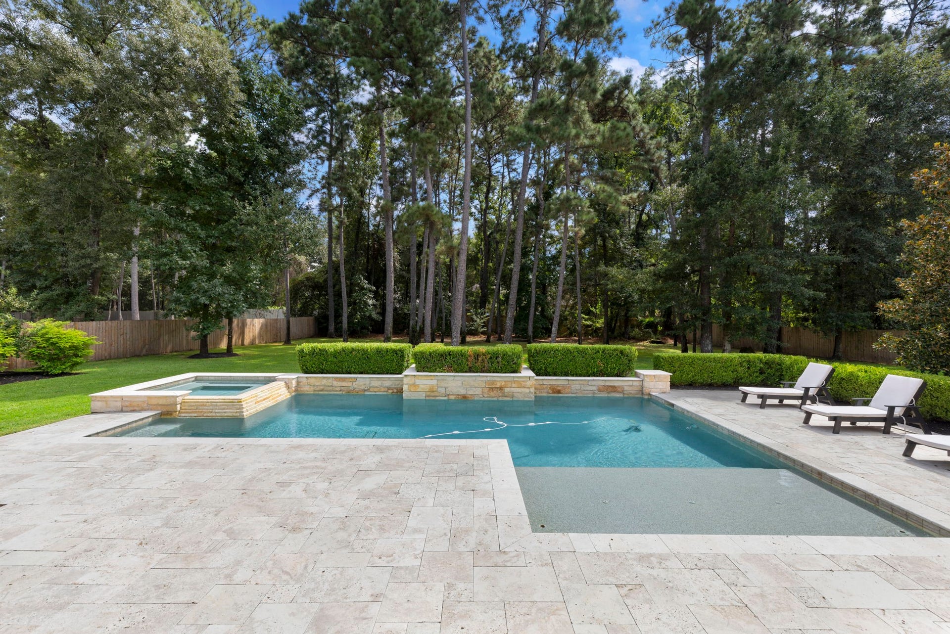 A sparkling geometric pool and raised spa are framed in natural stone coping, bordered by manicured hedges and a spacious travertine sundeck.