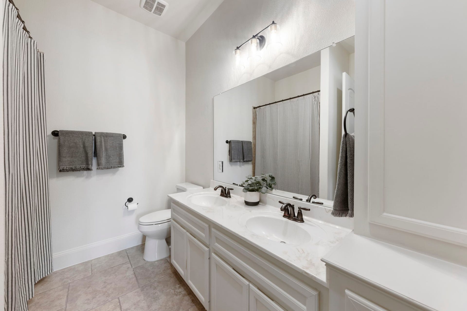 A generously sized shared bath offers a dual-sink vanity topped in marble-look stone and oil-rubbed bronze fixtures, finished with warm neutral tile flooring throughout.