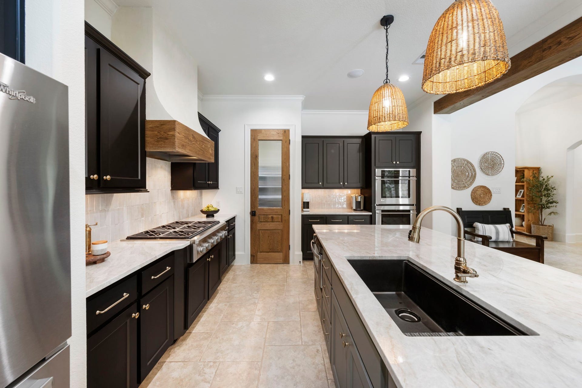 A functional prep galley pairs a commercial-style gas range with a stainless refrigerator, warm zellige backsplash, and a solid wood pantry door. An oversized workstation sink with a polished brass faucet anchors the adjoining island.