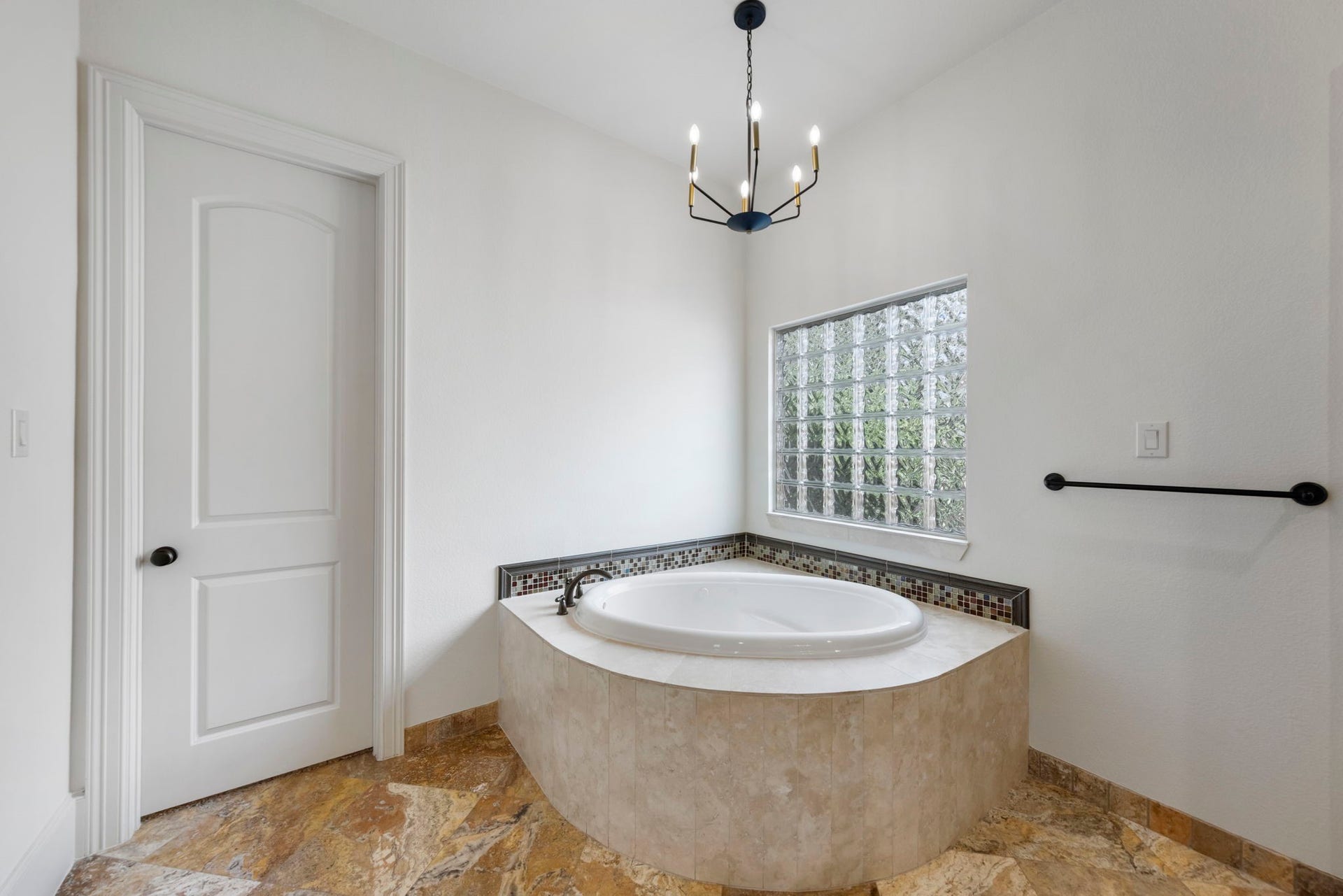 A generously sized corner soaking tub is framed in warm stone tile with a mosaic accent border, illuminated by a statement chandelier and glass-block window.