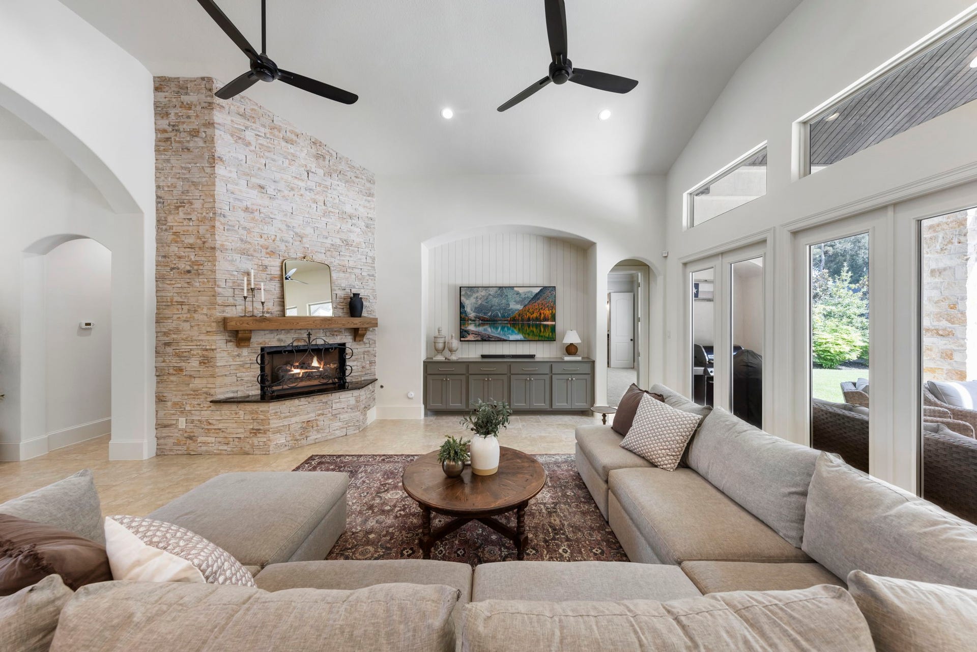 A dramatic stacked-stone fireplace with a rustic wood mantel serves as the centerpiece of this bright, airy living space, framed by oversized windows.