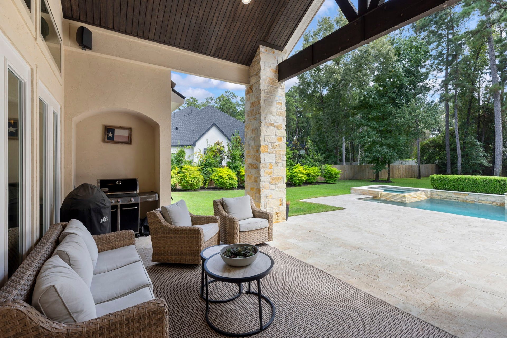 A vaulted wood-plank ceiling and dramatic timber trusses crown this expansive covered patio, complete with a built-in outdoor kitchen niche and stone columns overlooking the pool.