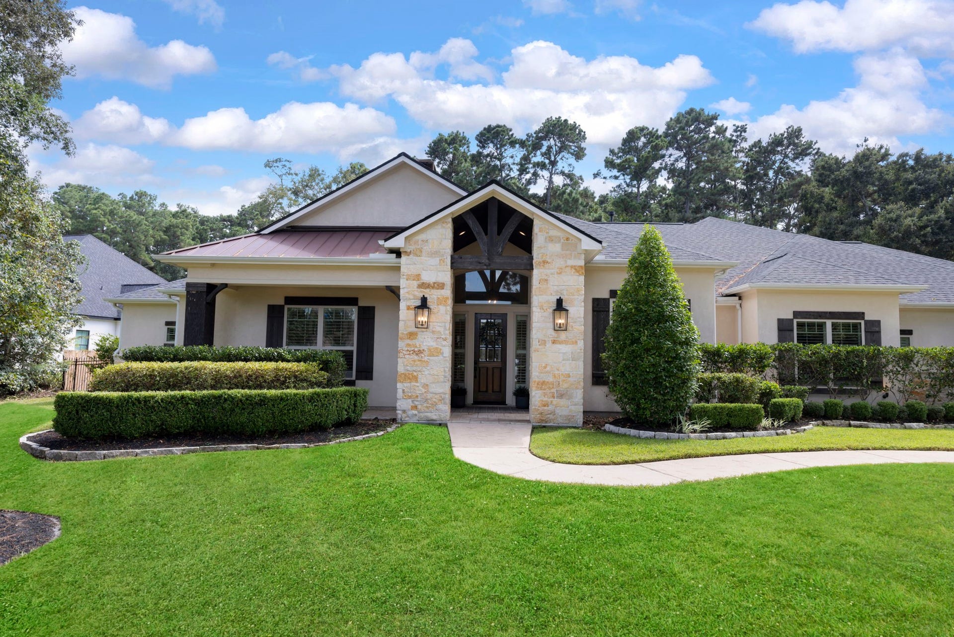 Charming stone-and-stucco elevation set behind a lushly manicured lawn, framed by mature pines and clipped boxwood hedges. A dramatic timber-and-stone entry gable and gracefully curving paver walkway deliver instant curb appeal.
