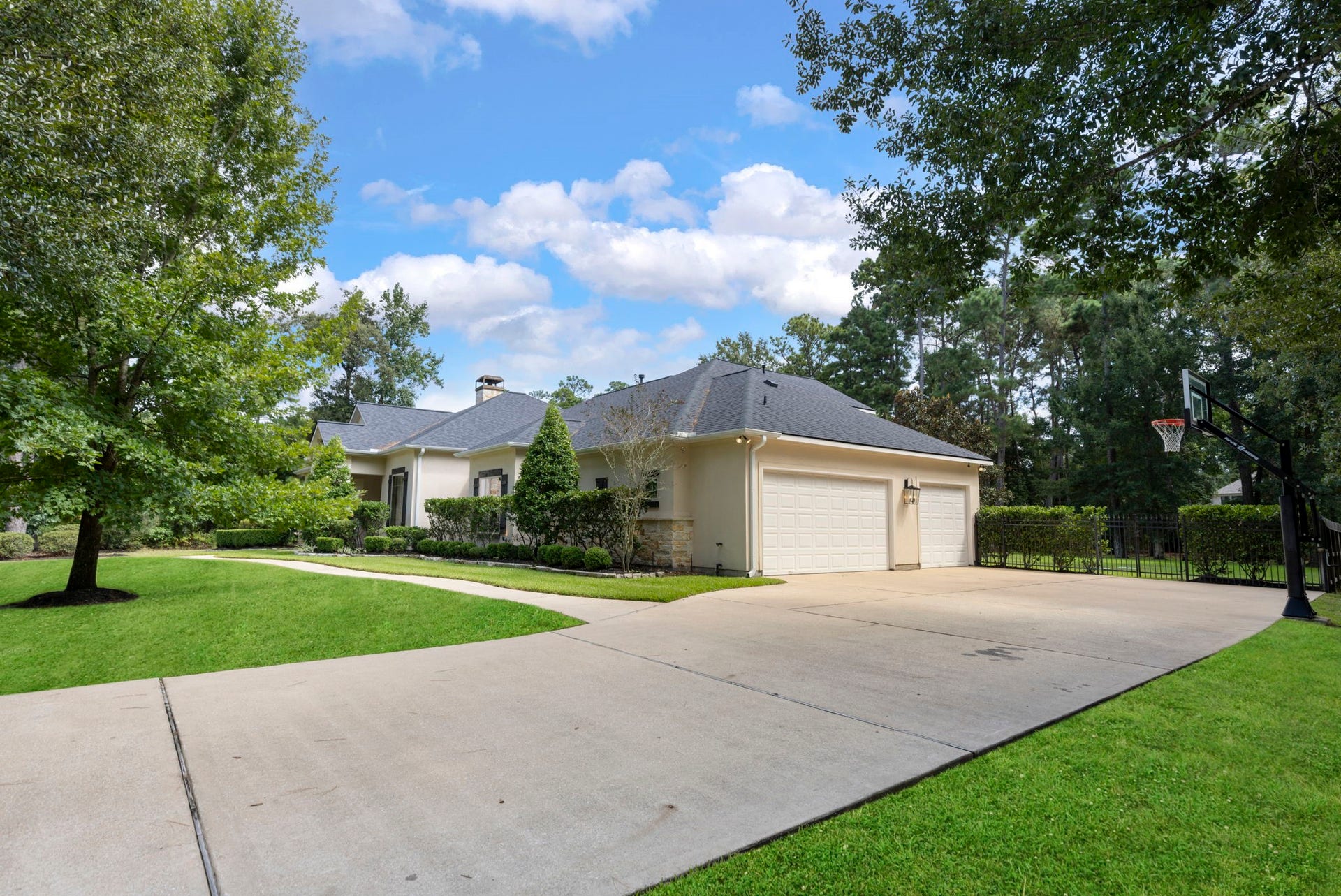 An expansive concrete driveway leads to the attached three-car garage, with a private basketball hoop and wrought-iron side gate hinting at the fenced backyard beyond. Towering shade trees and a sweeping side lawn add to the peaceful, wooded setting.