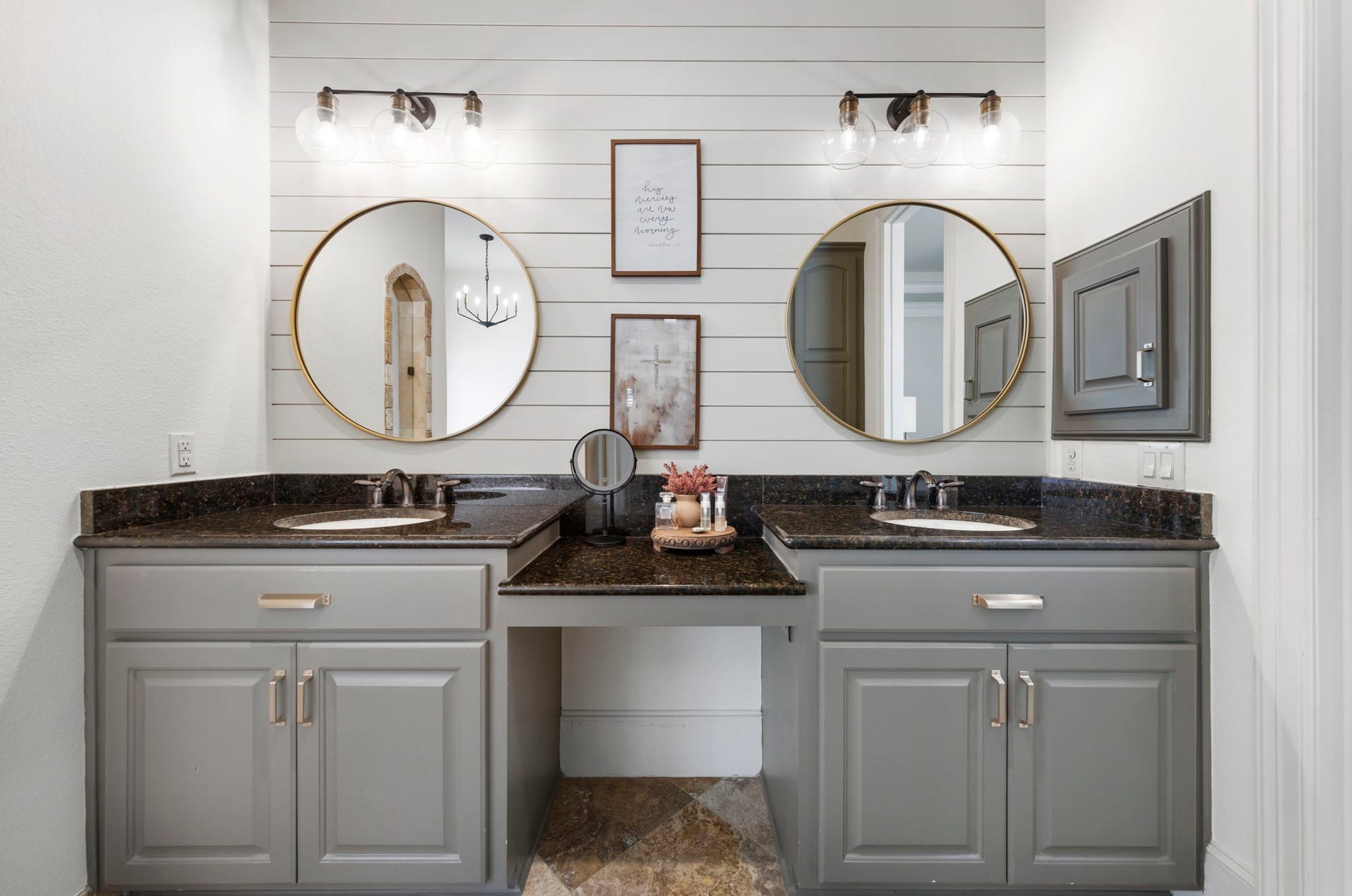 Dual vanities topped in rich granite are framed by oversized round mirrors and modern sconce lighting against a shiplap backdrop, offering his-and-hers convenience.