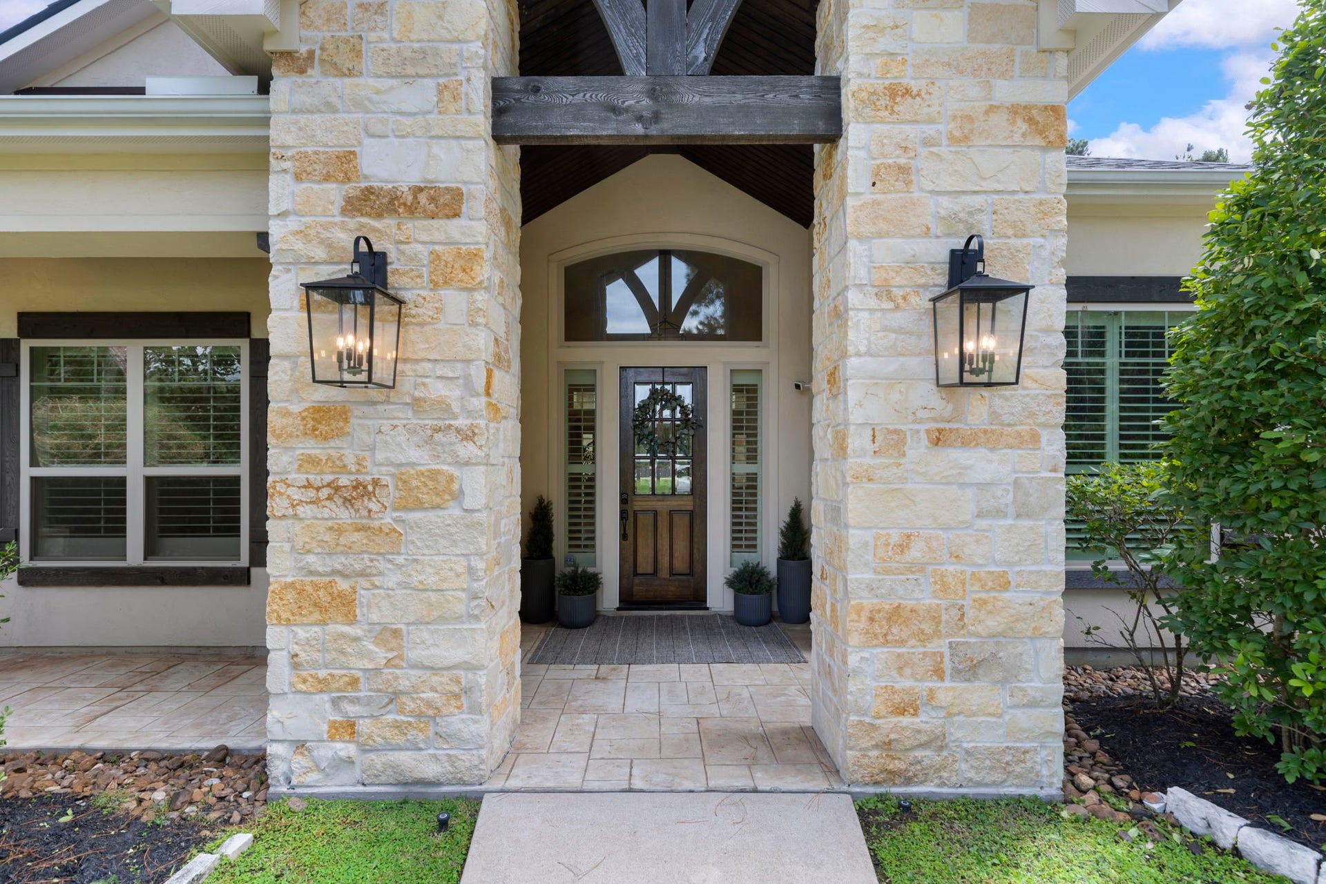 Hand-cut limestone columns and a rugged timber beam frame the arched glass transom above a rich wood front door dressed with a seasonal wreath. Warm lantern sconces and potted greenery lend refined, welcoming charm.