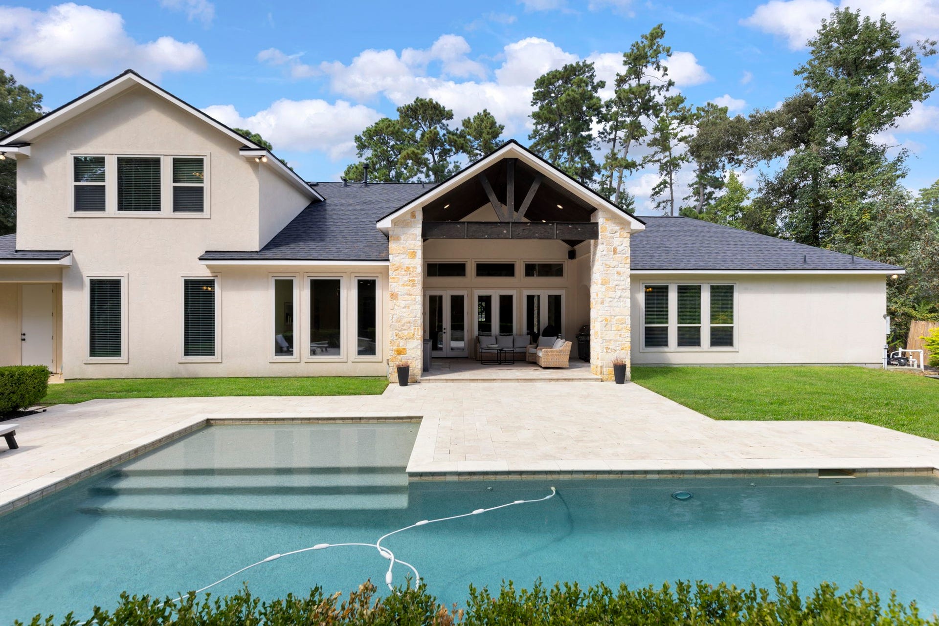 The home's back elevation showcases soaring stucco gables, a dramatic timber-frame covered porch, and walls of windows overlooking the resort-style backyard.