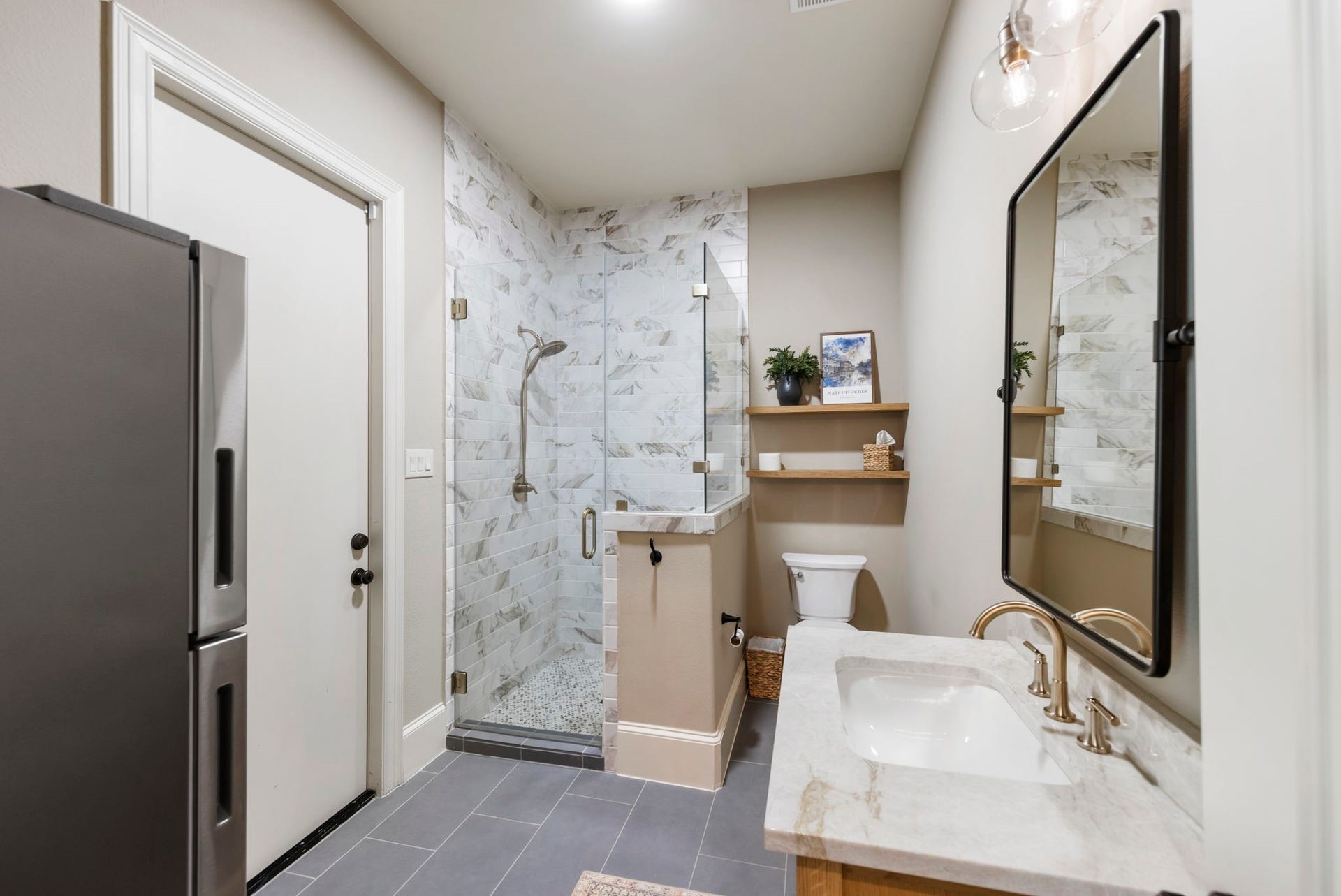 Sleek marble-look subway tile lines the walk-in shower, paired with a half-wall tile surround and warm brass fixtures. Doubling as a convenient pool bath, it offers easy access for guests coming in from the backyard, with open wood shelving for handy storage.