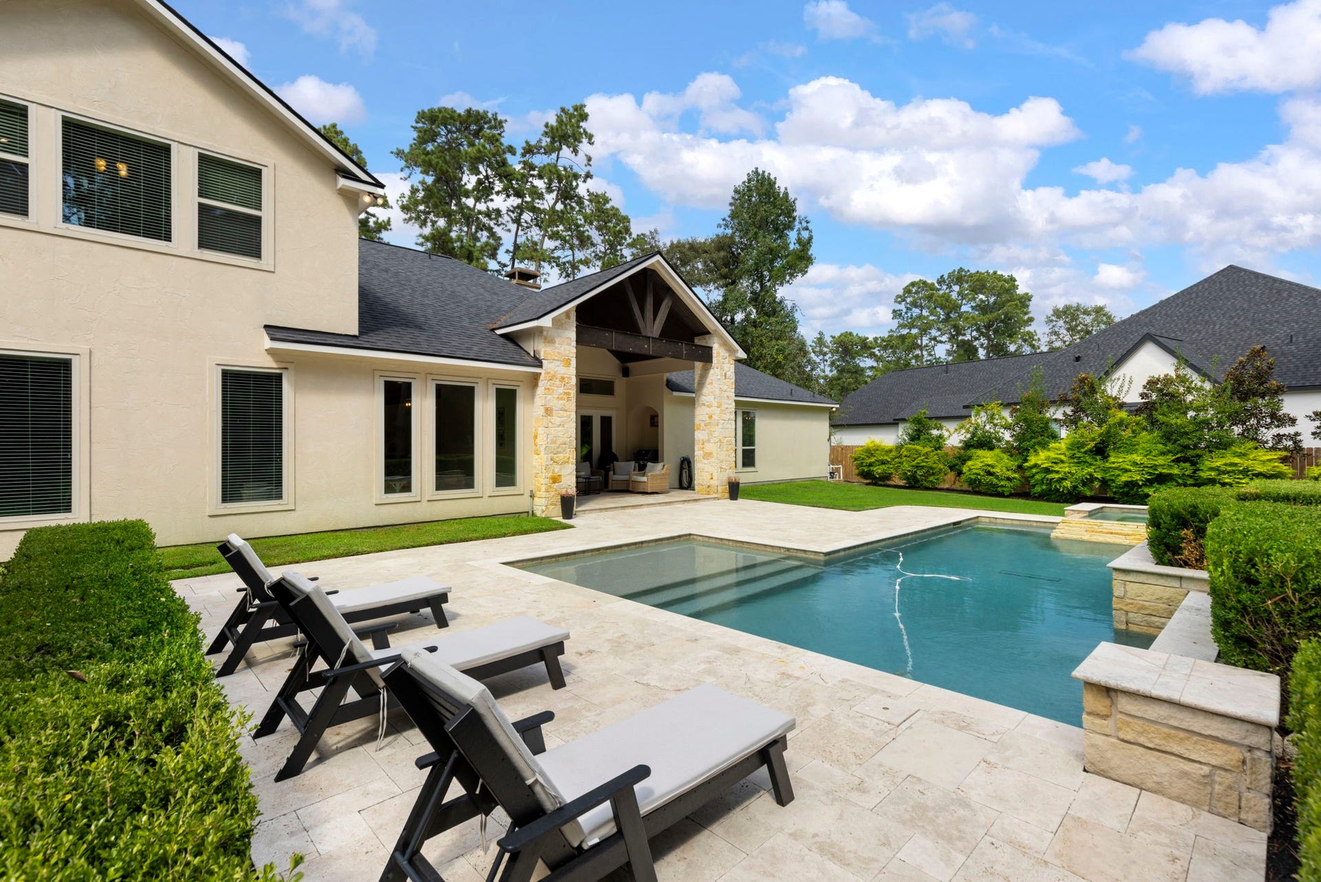 A stately two-story back elevation rises beyond the sun-splashed travertine patio, geometric pool, and raised spa, all framed by lush, mature landscaping.