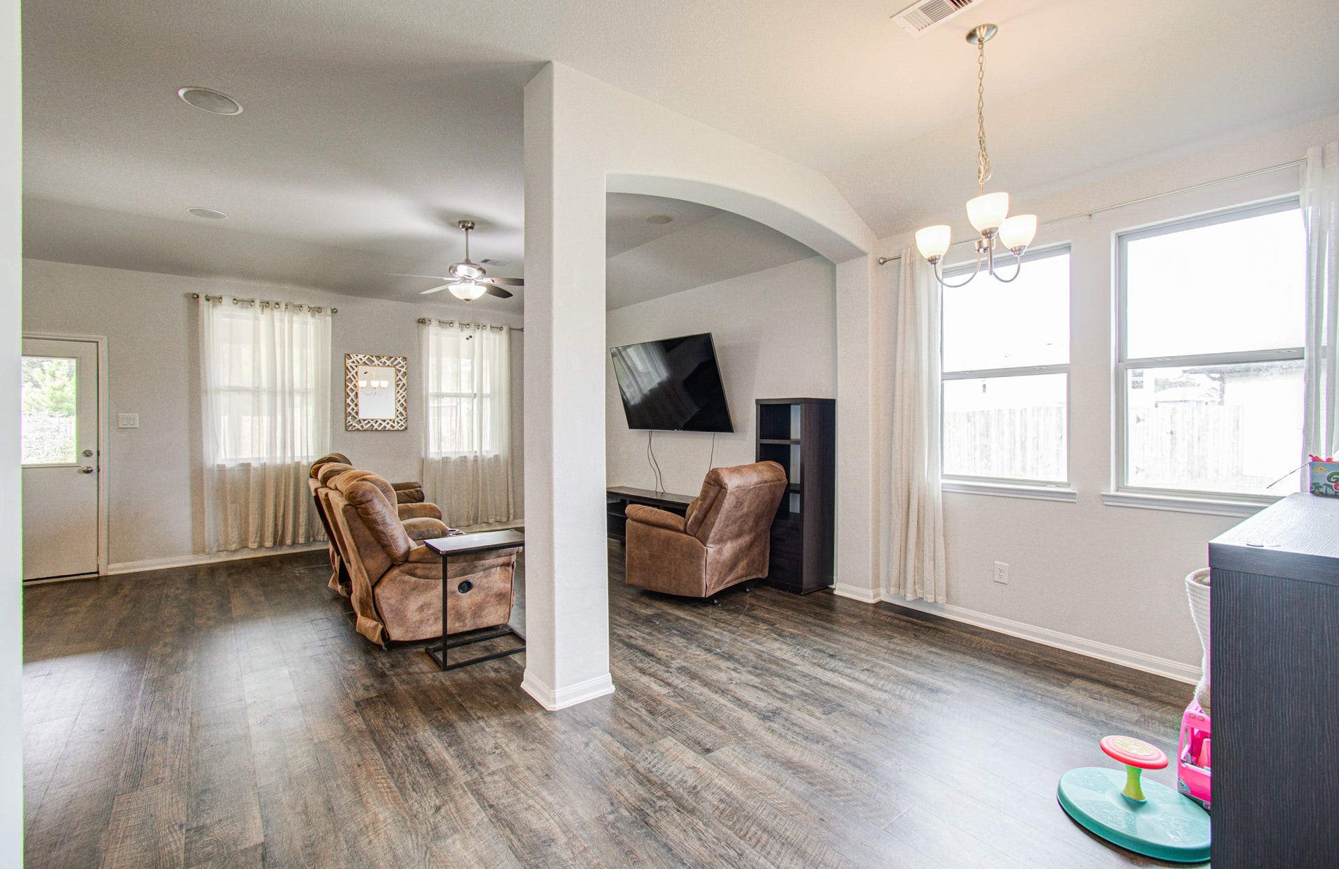 11064 Patriot Court, Cleveland, TX 77328 Bright and spacious living area featuring elegant wood-like flooring, cozy recliners, and a wall-mounted TV, seamlessly flowing into a dining nook with large windows and a charming chandelier.