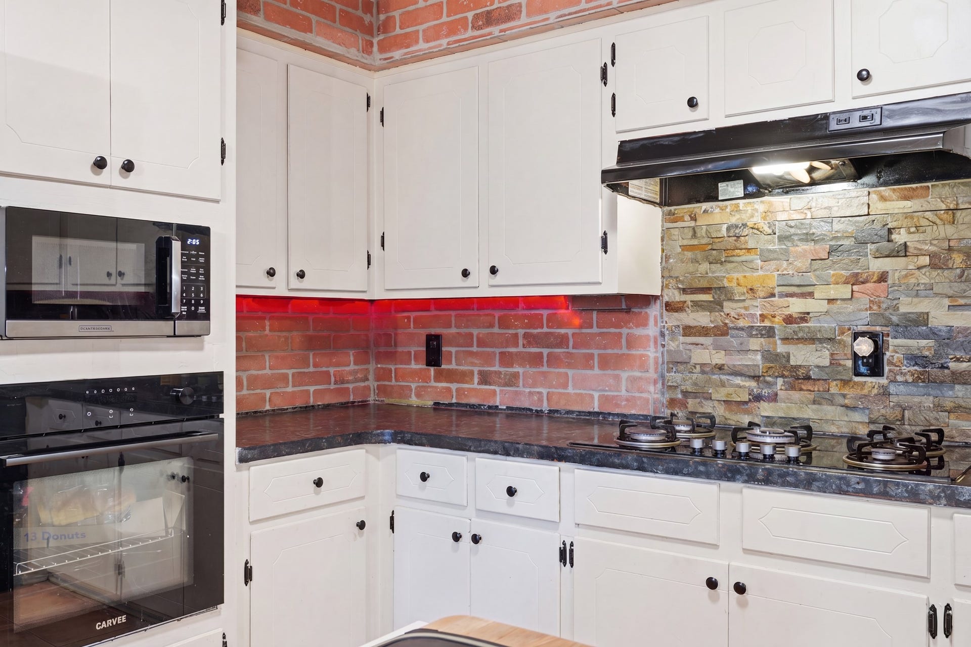 2216 Peach Lane, Pasadena, TX 77502 This kitchen features a modern design with white cabinetry, sleek black appliances, and a stylish mix of red brick and stone backsplash. The dark countertops add contrast, and there's under-cabinet lighting for a warm ambiance.