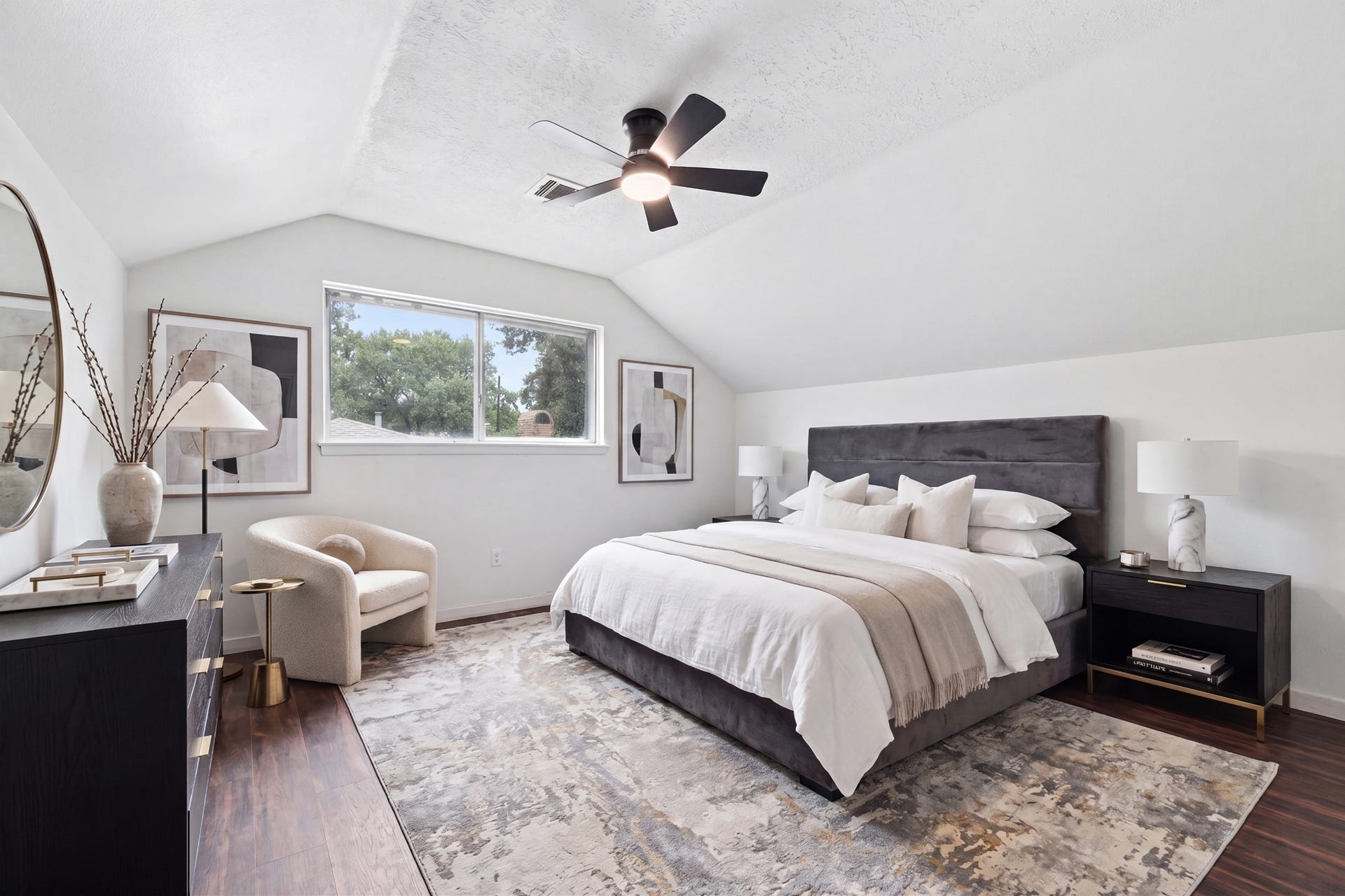 2216 Peach Lane, Pasadena, TX 77502 This bedroom features a vaulted ceiling with a modern fan, a large window offering natural light, and a cozy, neutral color palette. It includes a plush bed with ample pillows, stylish nightstands with lamps, and a comfortable armchair. The room is accented with tasteful decor and a soft area rug on hardwood floors.