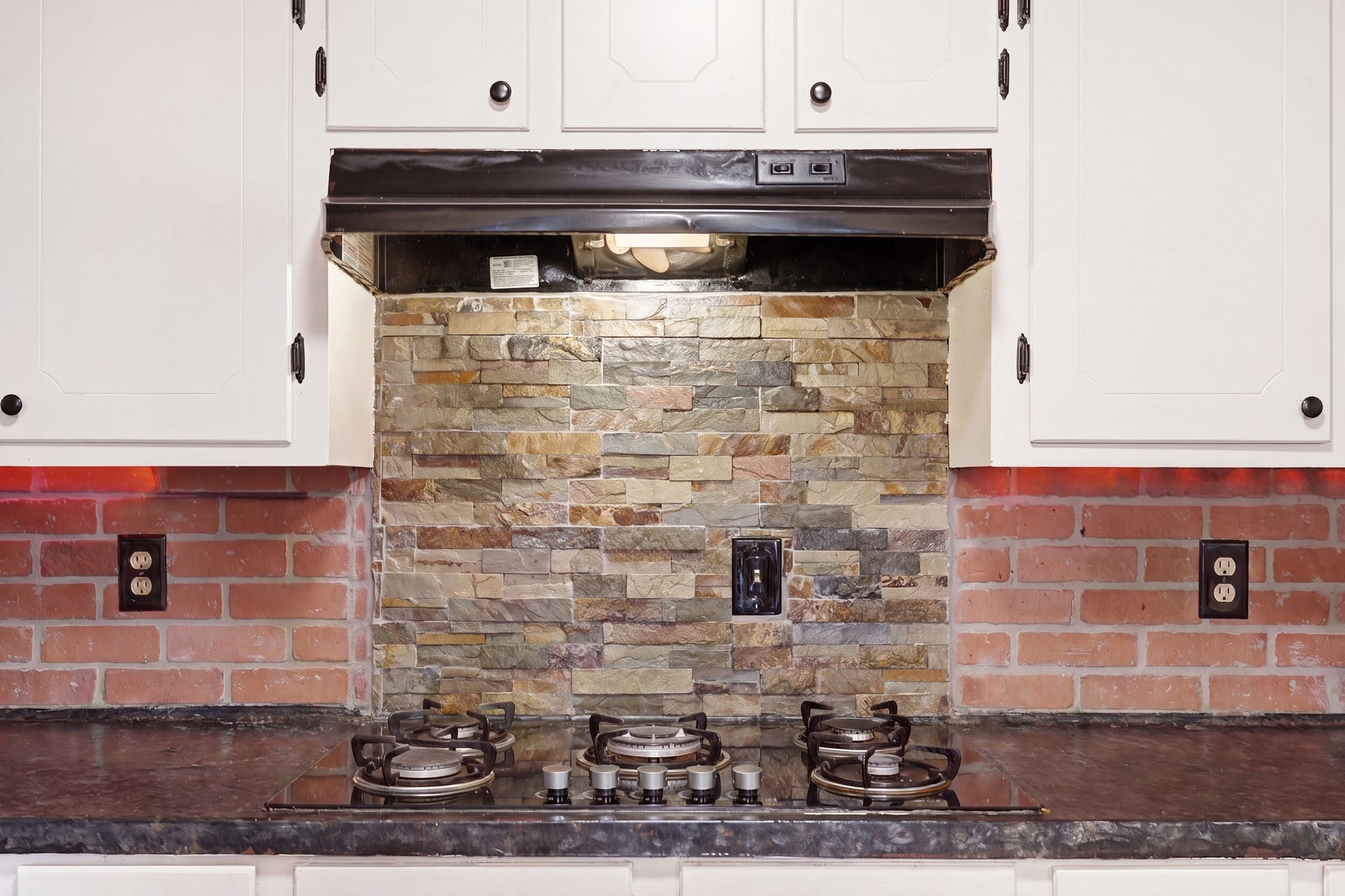 2216 Peach Lane, Pasadena, TX 77502 This kitchen features a stylish backsplash with a mix of brick and stone tiles, complemented by white cabinetry and a modern black cooktop.