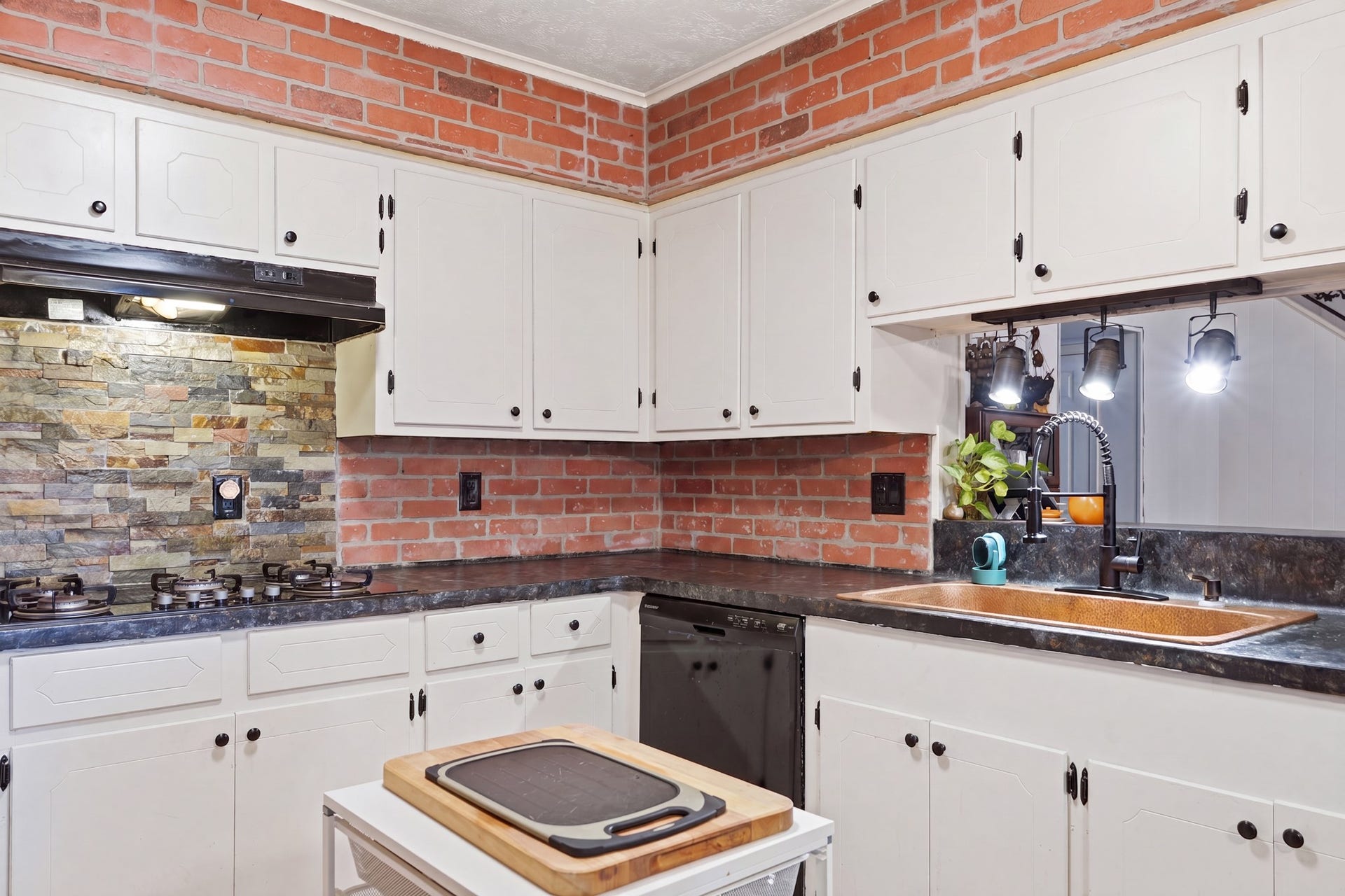 2216 Peach Lane, Pasadena, TX 77502 This kitchen features a cozy, rustic design with exposed brick walls and a stone backsplash. It has white cabinetry, a modern sink with stylish lighting, and a built-in cooktop, offering both functionality and charm.