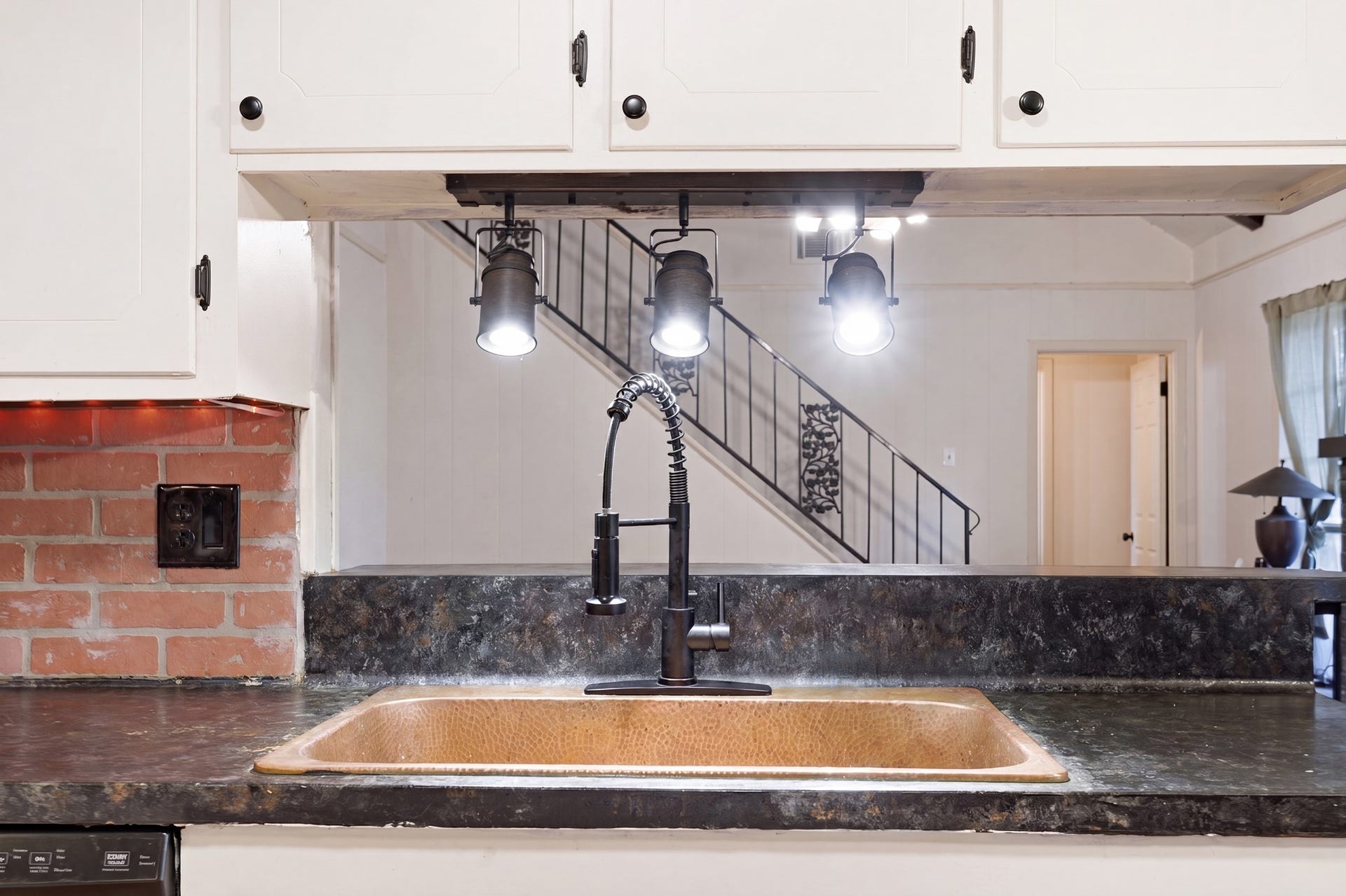 2216 Peach Lane, Pasadena, TX 77502 This kitchen features a stylish copper sink with a modern black faucet, set against a dark countertop. White cabinetry and a brick backsplash add contrast, while industrial-style pendant lights provide illumination. A staircase is visible in the background, adding architectural interest.